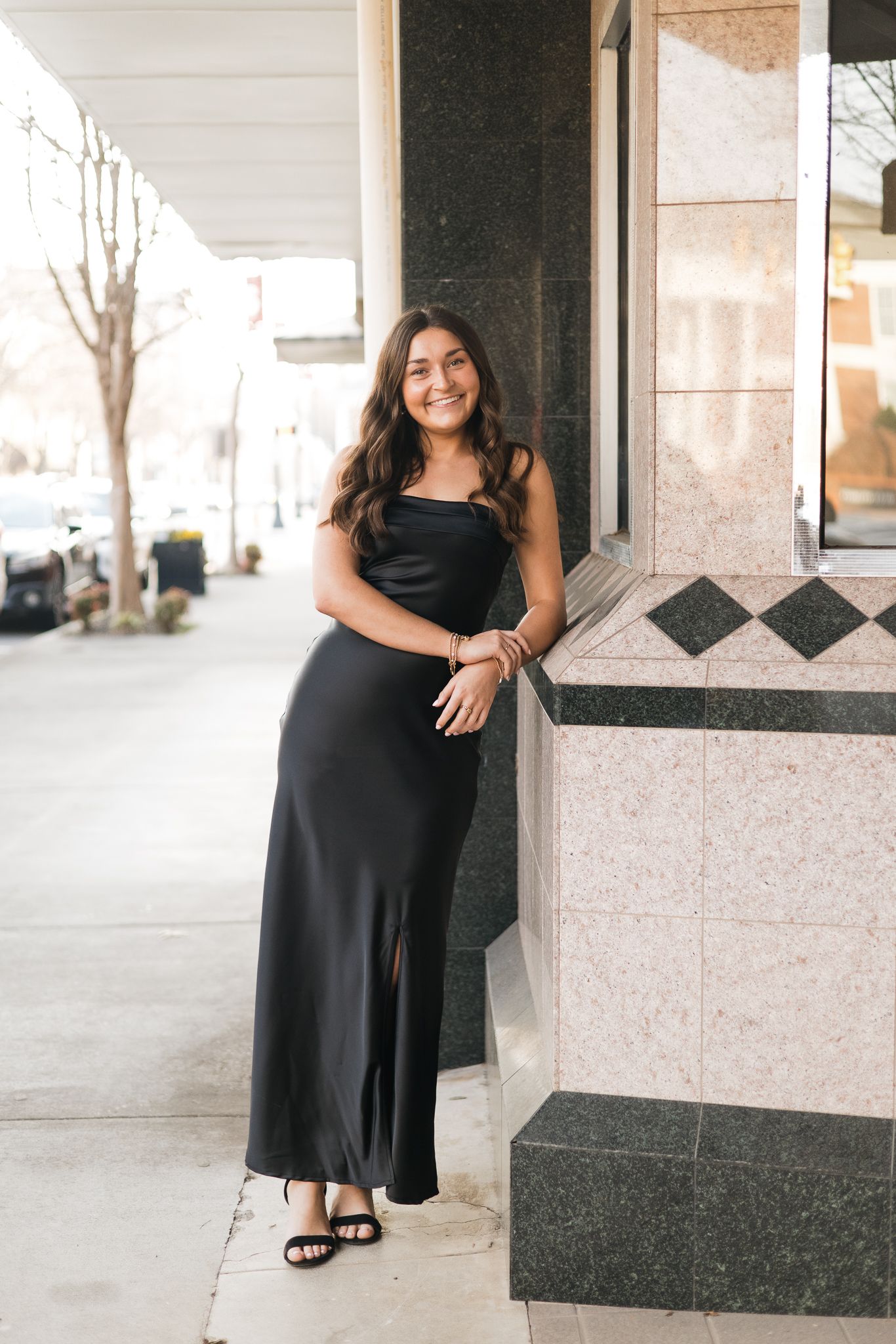 Woman in black maxi slip dress with slit and heeled sandals posing against a marble column on a city sidewalk.