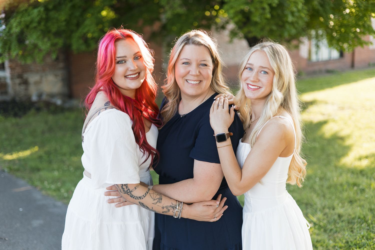 Three women smiling and posing outdoors, two in white dresses and one in navy, surrounded by green grass and trees.