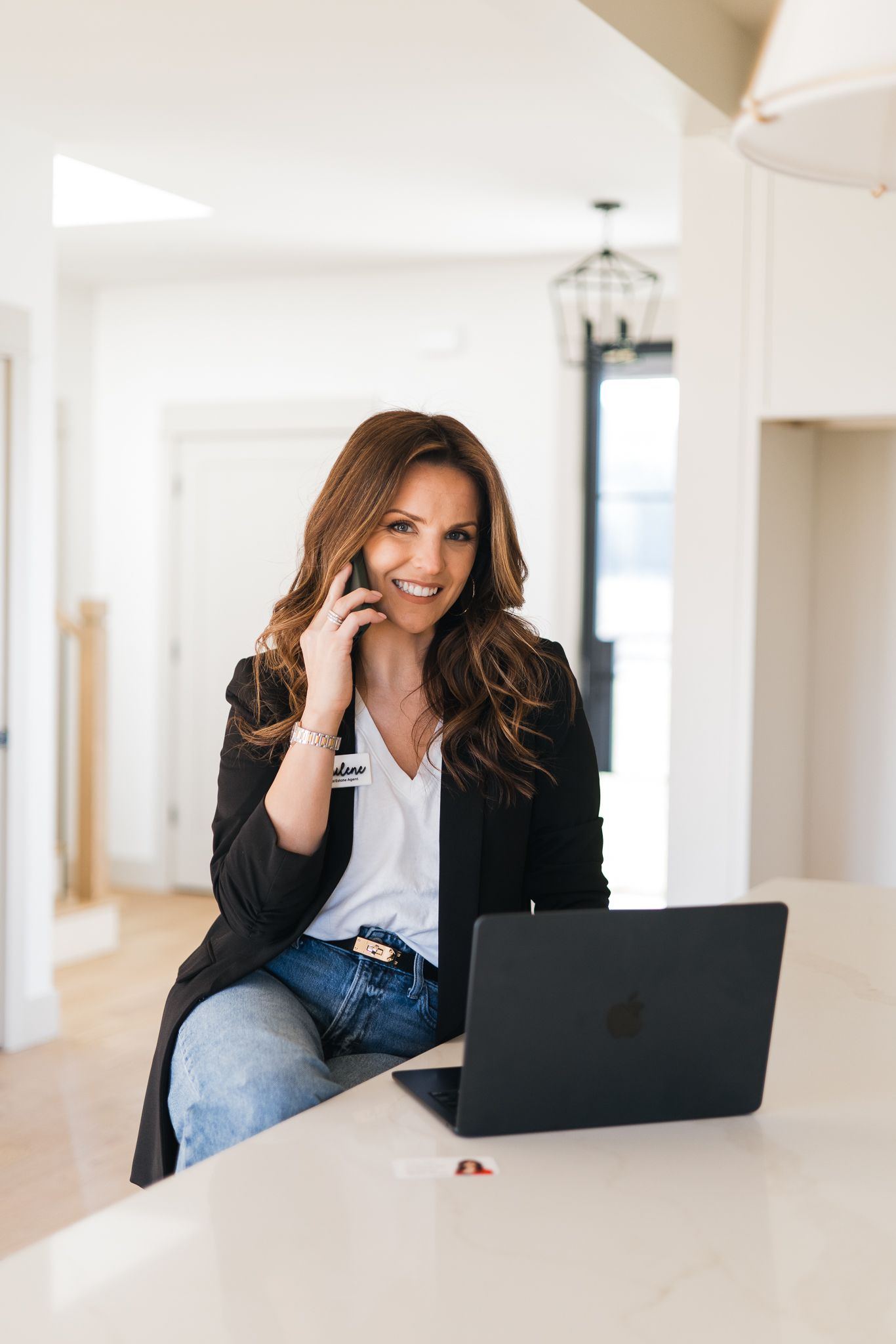 Smiling brunette woman in black blazer on phone with laptop at a bright modern kitchen counter.