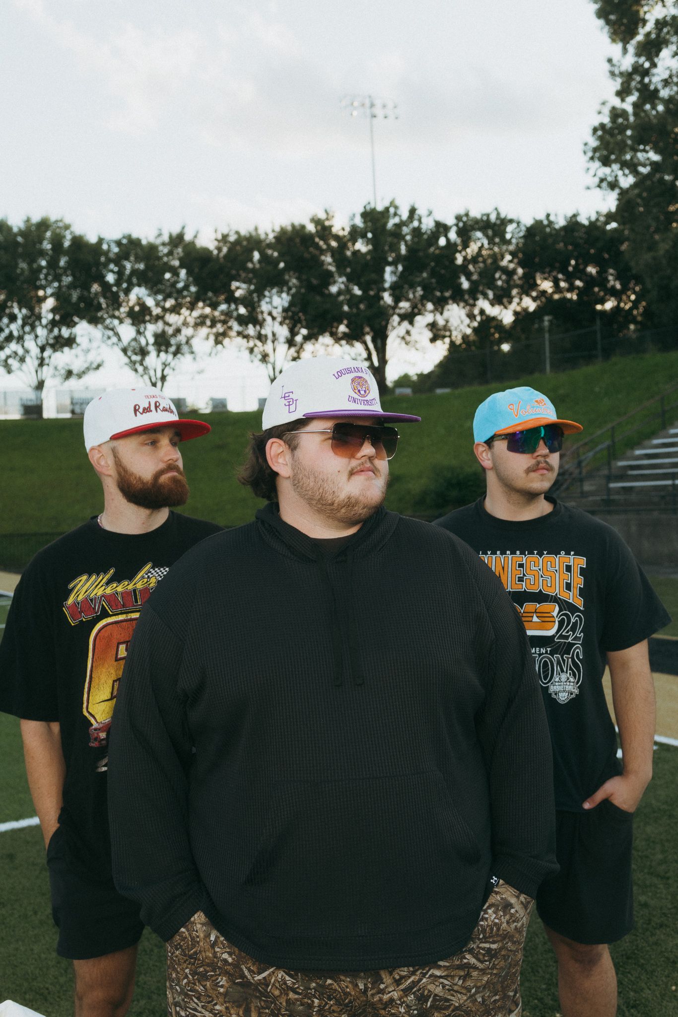 Three men wearing snapback caps posing on a football field at dusk.