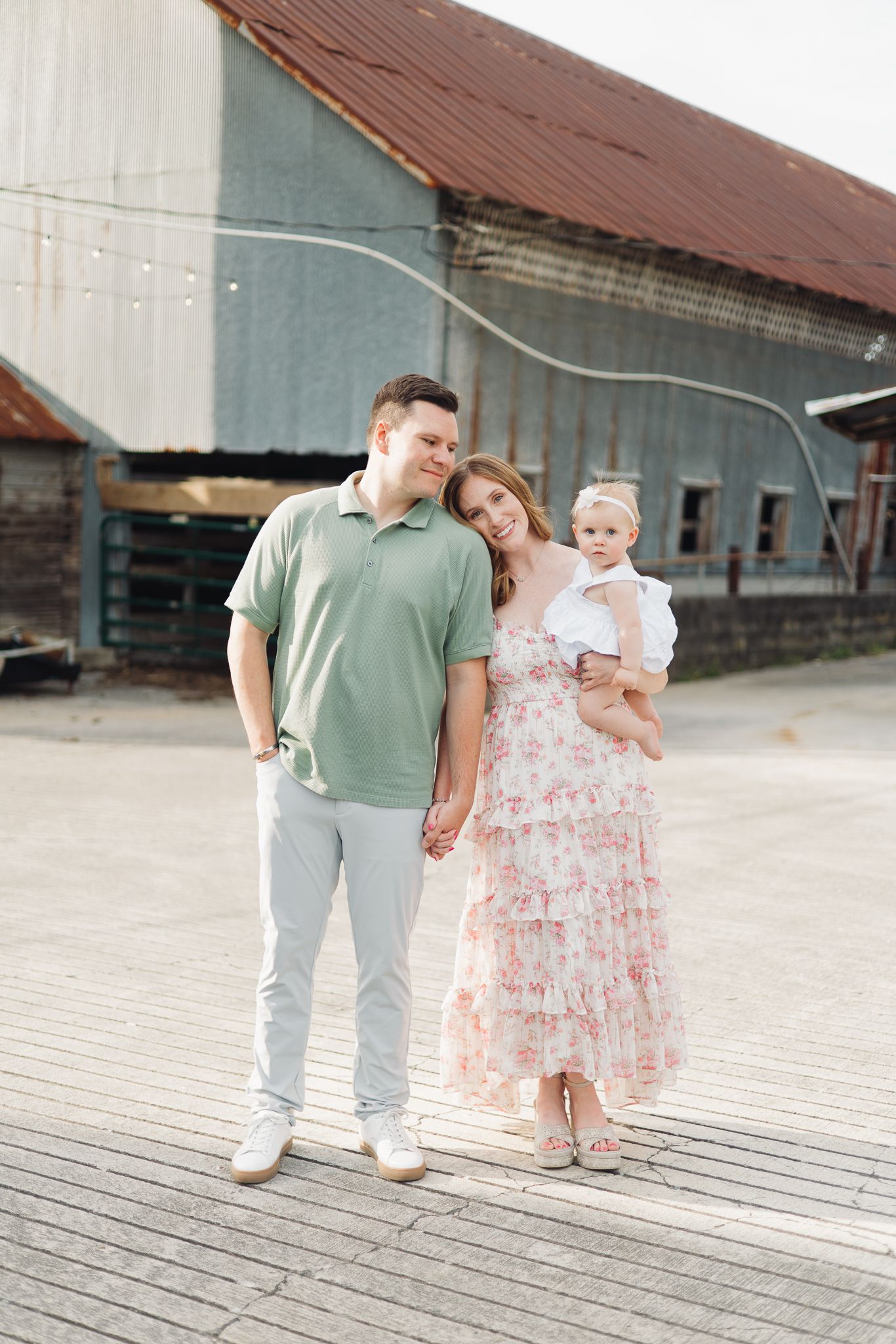 A family of three poses in front of a rustic barn, parents holding hands with mom cradling their baby girl.