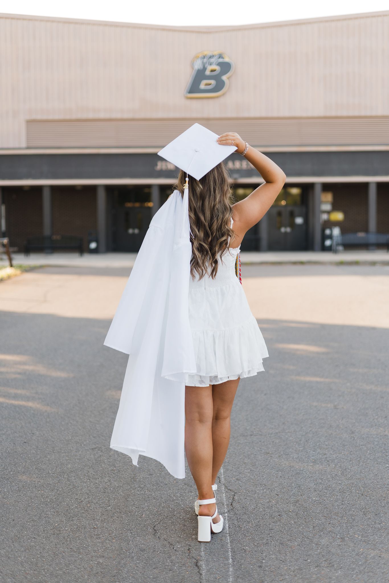 High school senior in white graduation cap and gown poses from behind in front of school building.
