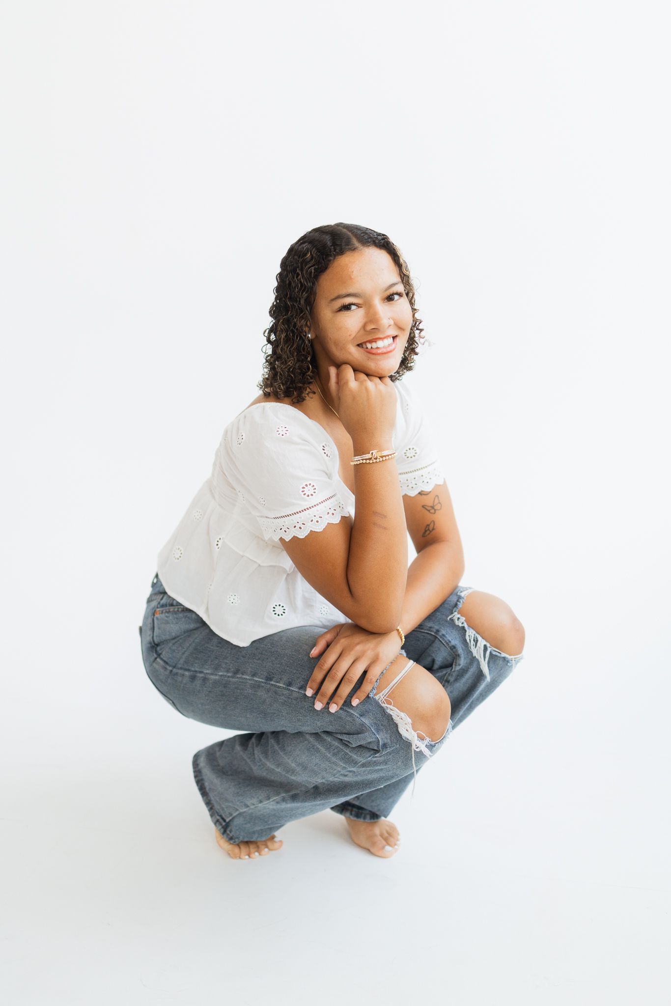 Smiling woman in white eyelet top and ripped jeans crouching barefoot against white background.