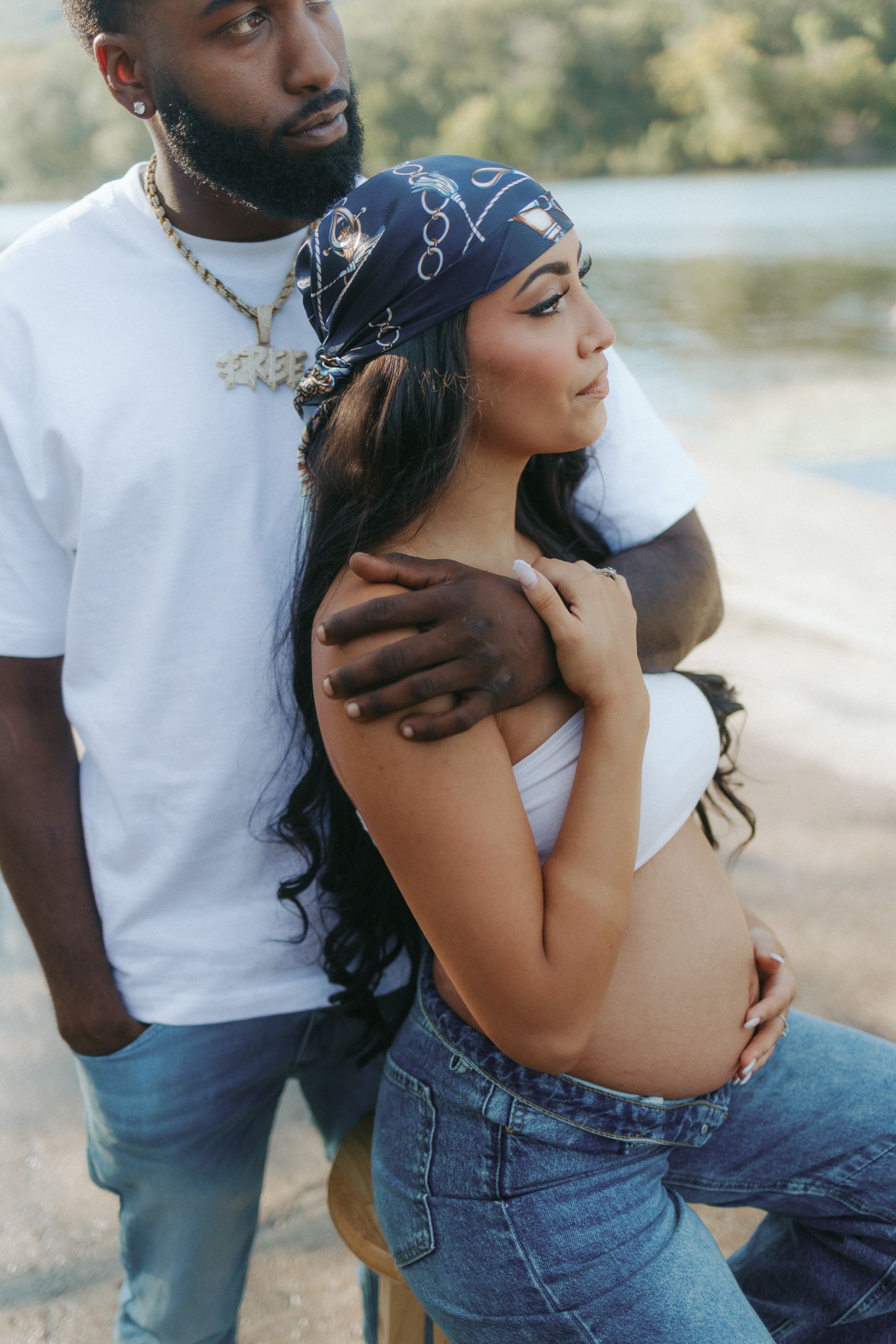 Pregnant woman cradling baby bump with partner embracing her shoulders near scenic waterside.