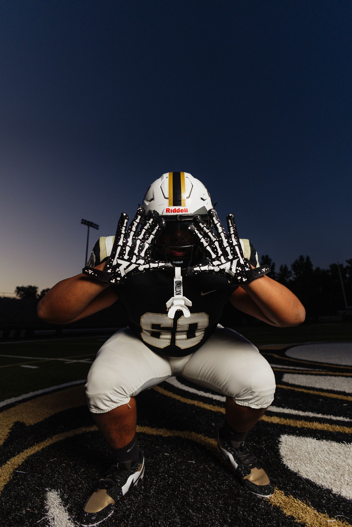 Football player in black jersey and white helmet crouches in defensive stance on field at dusk.