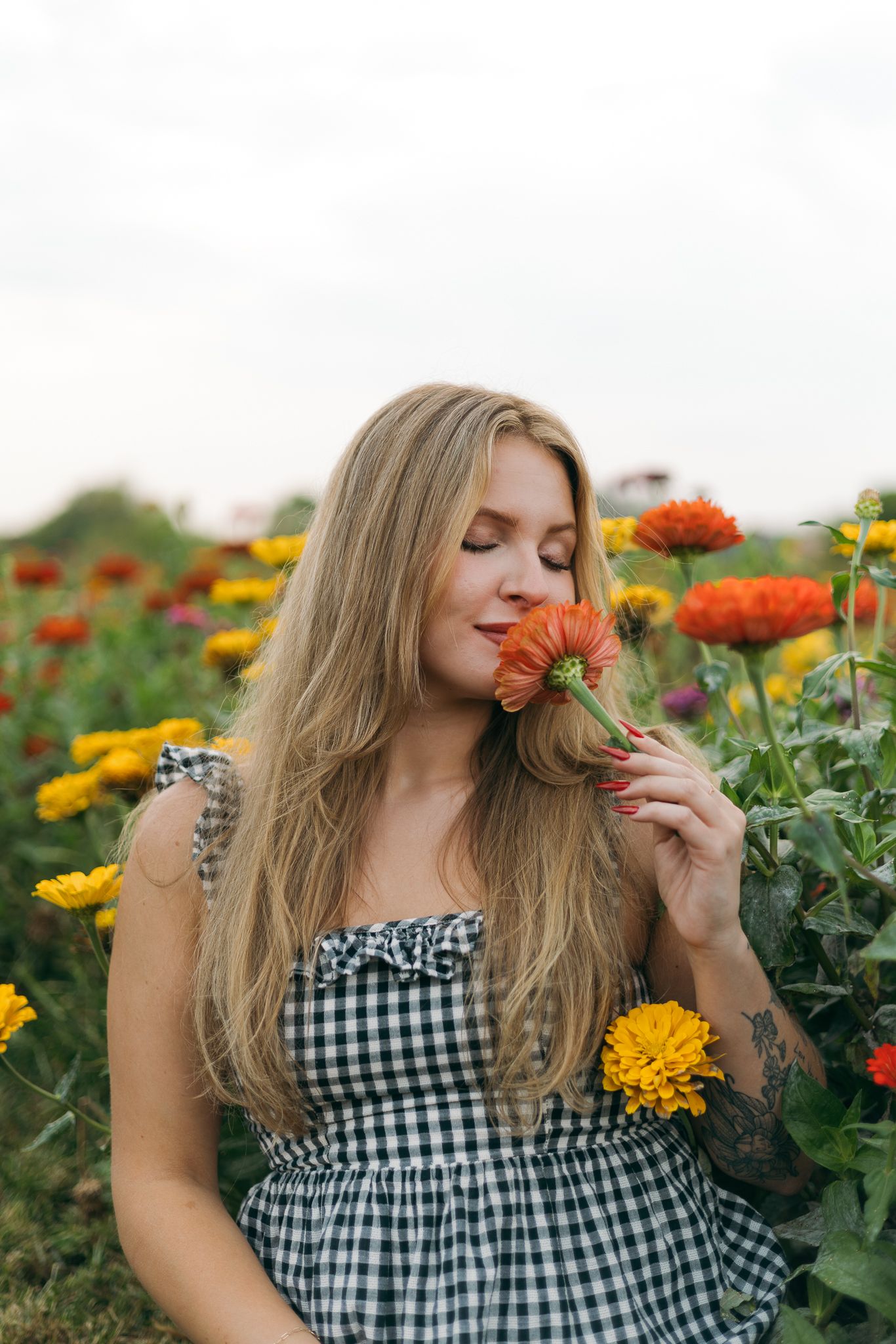 Blonde woman smelling orange zinnia flower in colorful flower field wearing gingham dress.