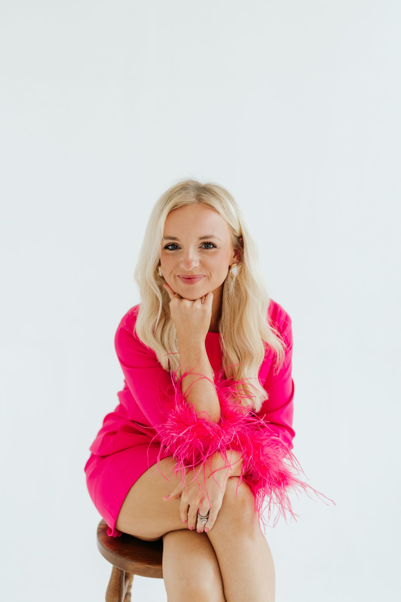 Smiling blonde woman in hot pink feather-trim dress sits on stool with chin resting on hand.
