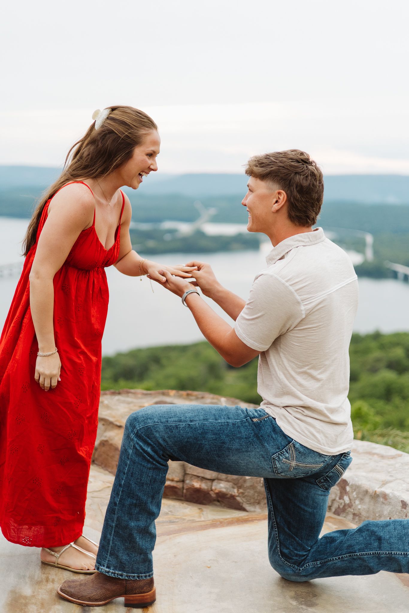 Man proposes on one knee to woman in red dress at scenic overlook with river and mountains in background.