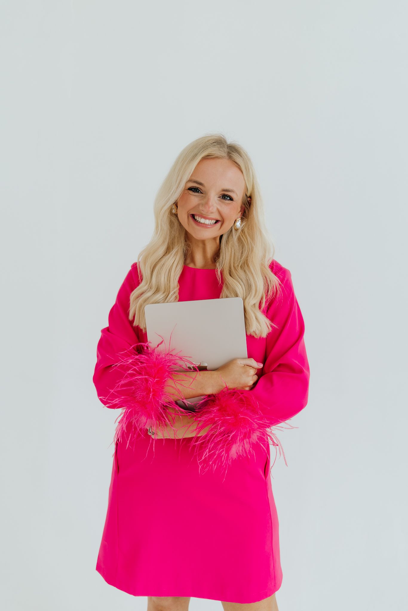Smiling blonde woman in pink feather-trim dress holds a laptop against white background.
