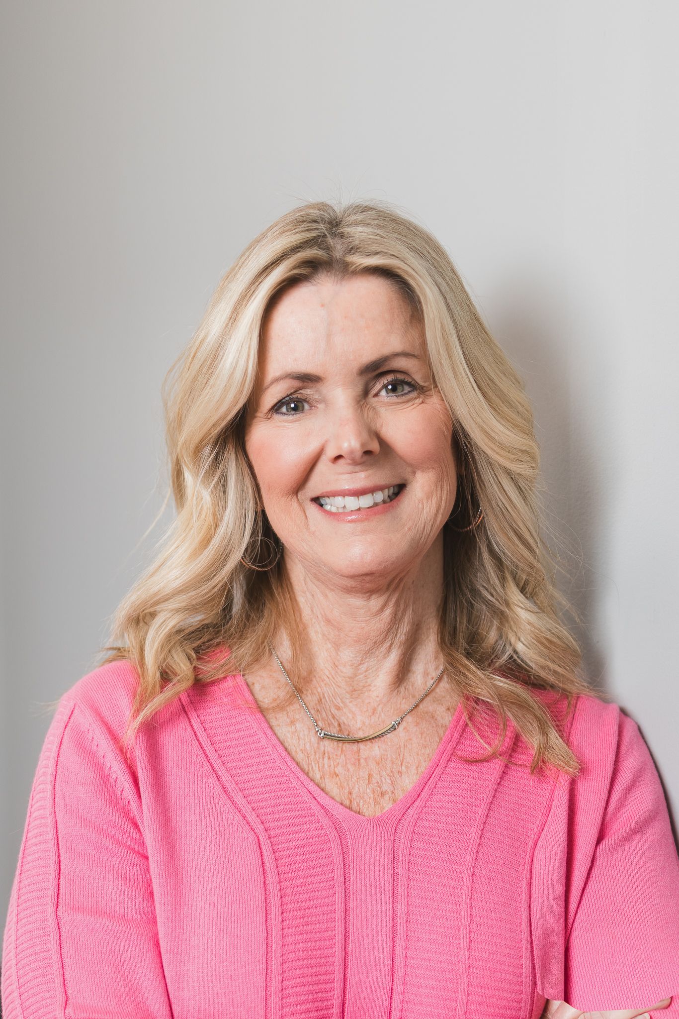 Smiling mature blonde woman in pink sweater posing against a neutral gray background.