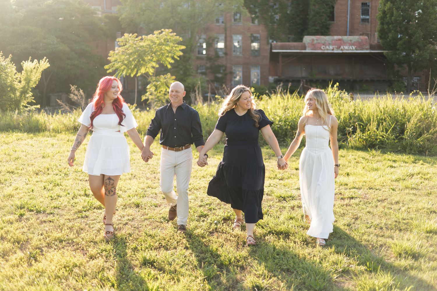 Happy family of four holding hands walking through sunny green field in coordinated outfits.