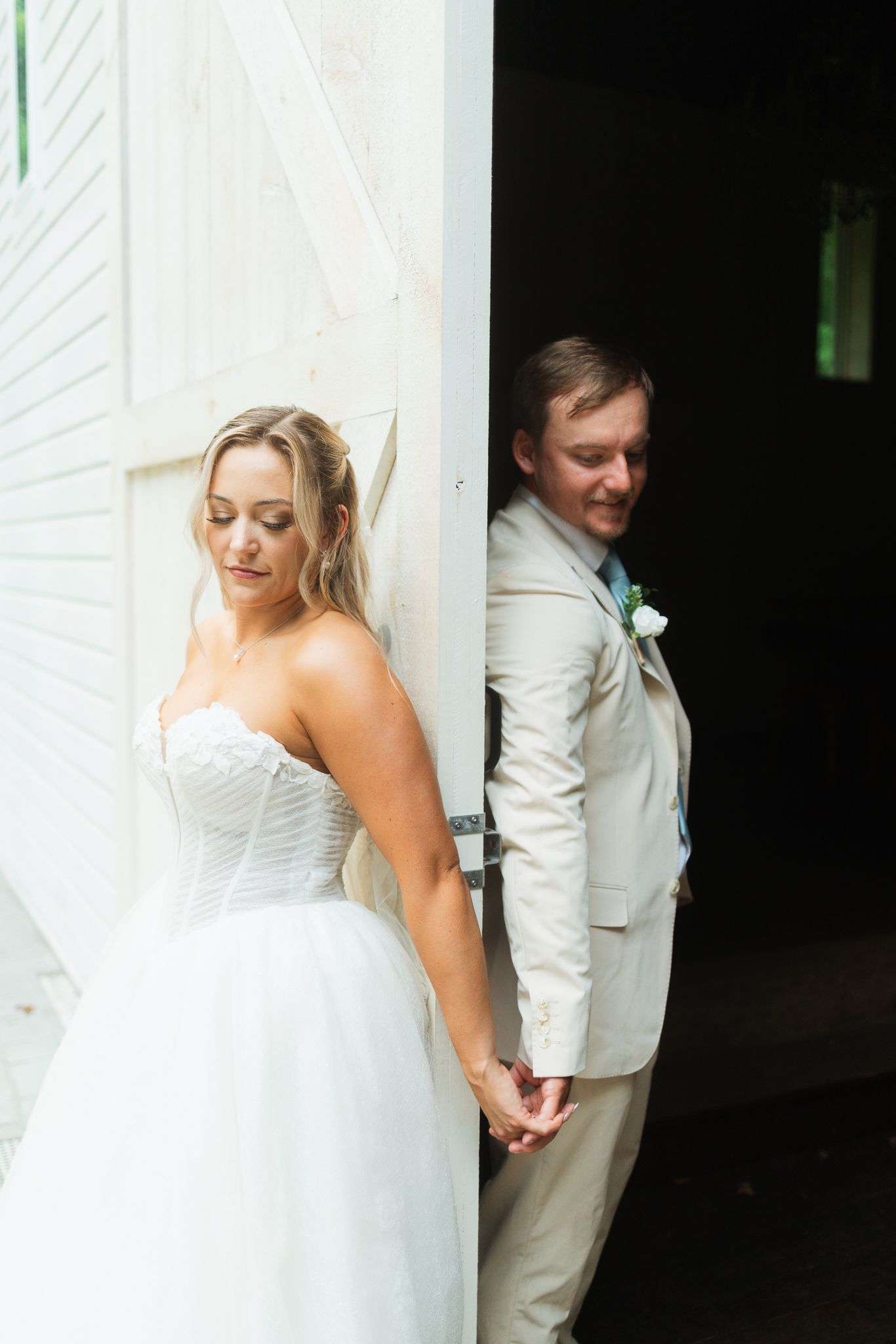 Bride in white strapless gown and groom in tan suit hold hands around a white barn door on their wedding day.
