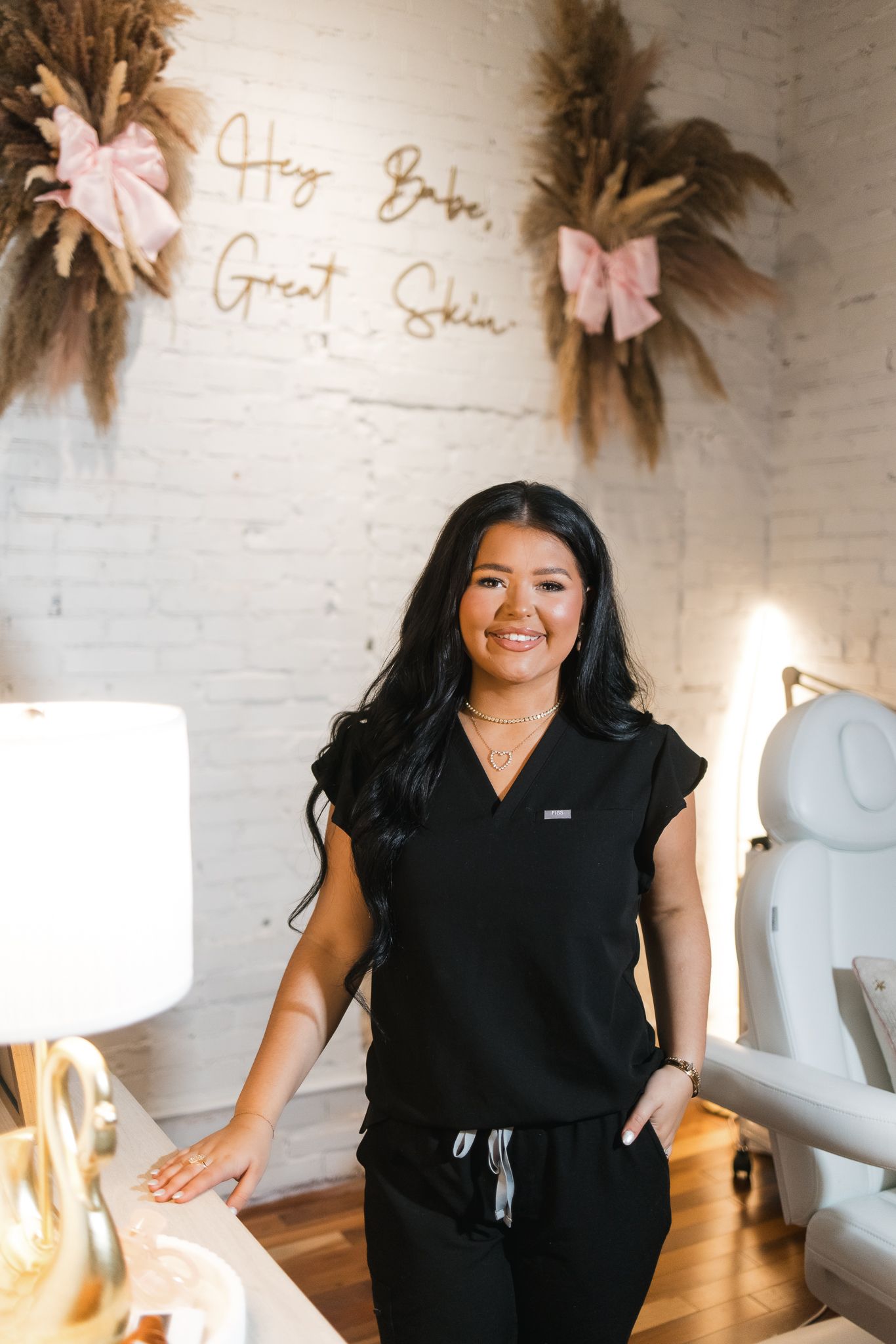 Smiling esthetician in black scrubs poses in a chic salon with a Hey Babe Great Skin neon sign.