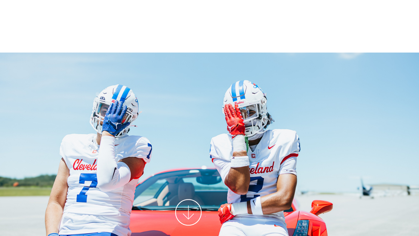 Two football players in blue and white uniforms conversing beside a red sports car on a runway.