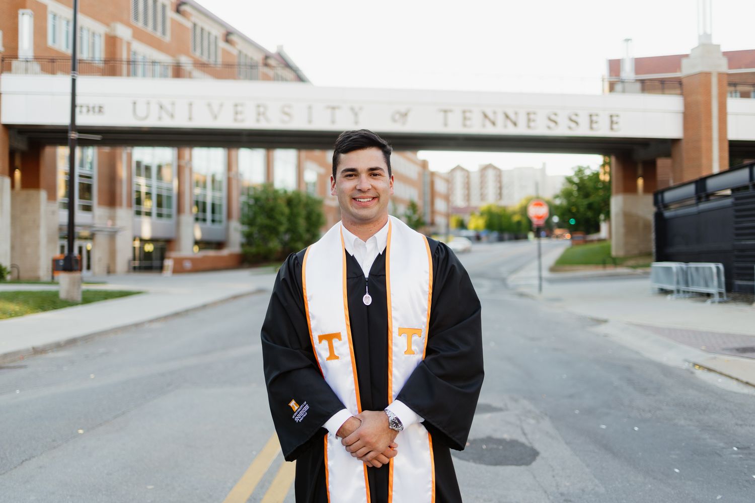 University of Tennessee graduate smiles in black cap and gown with orange and white stole in front of campus arch.