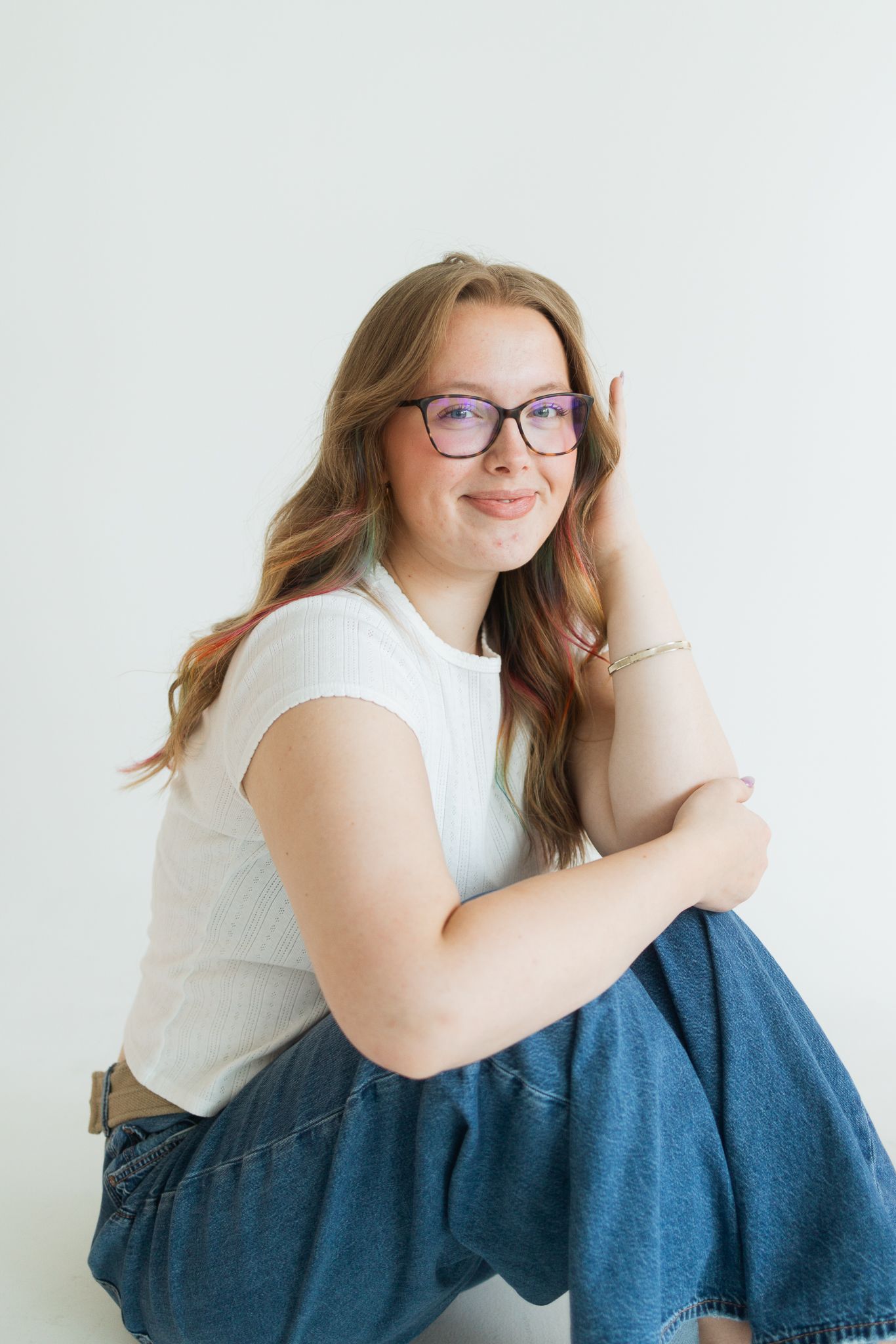 Young woman with glasses and long hair sits casually in jeans and white tee, smiling against white background.