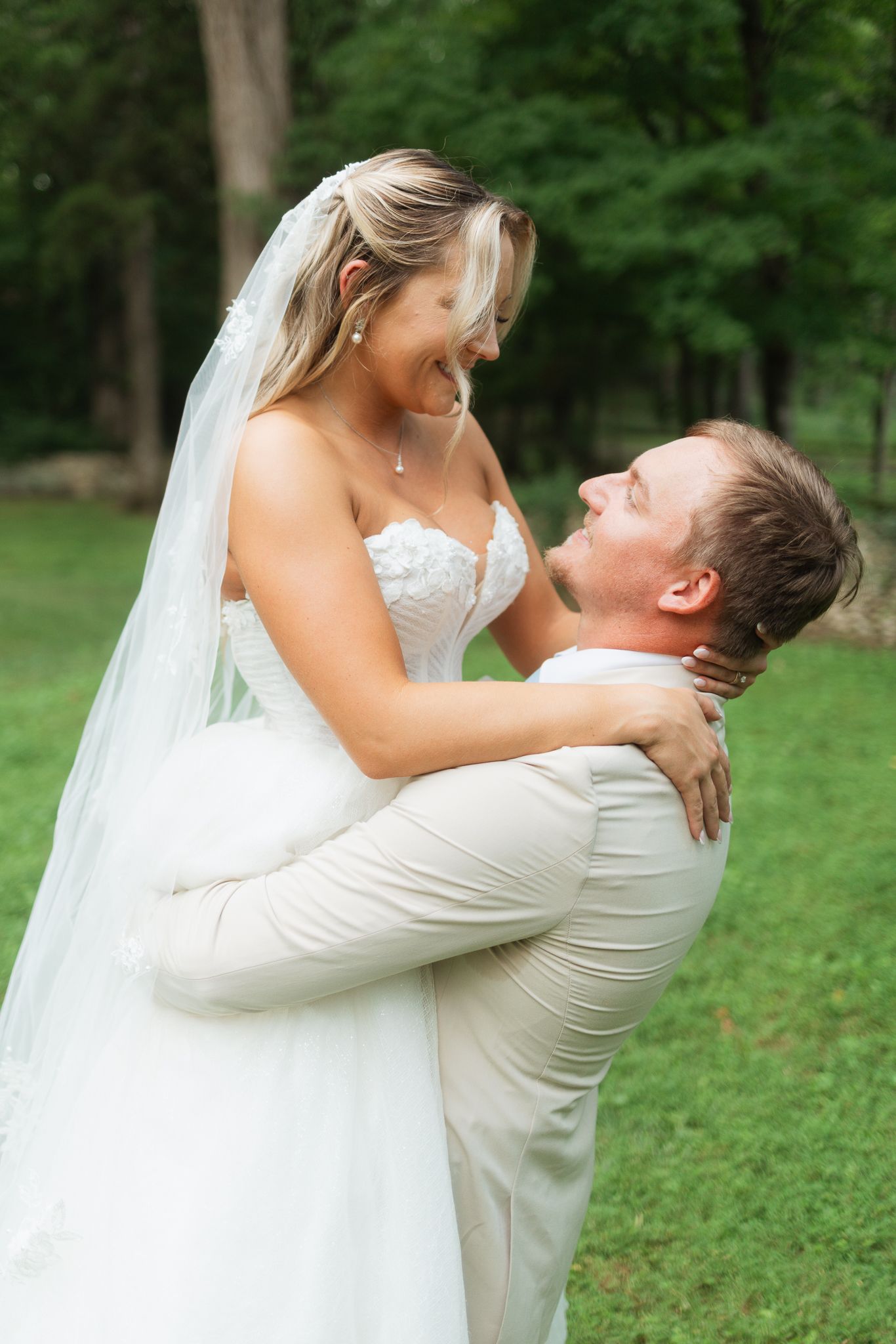 Groom lifting bride in white gown and veil, sharing a joyful moment outdoors in a lush green park setting.