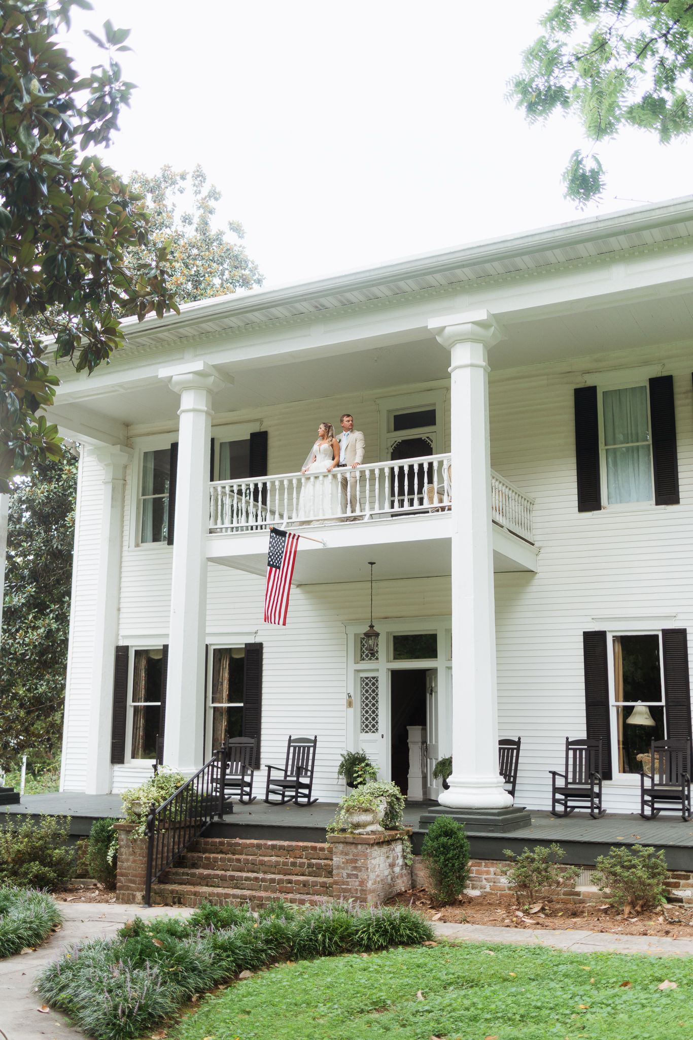 Bride and groom stand on balcony of white Southern colonial mansion with large columns and American flag.