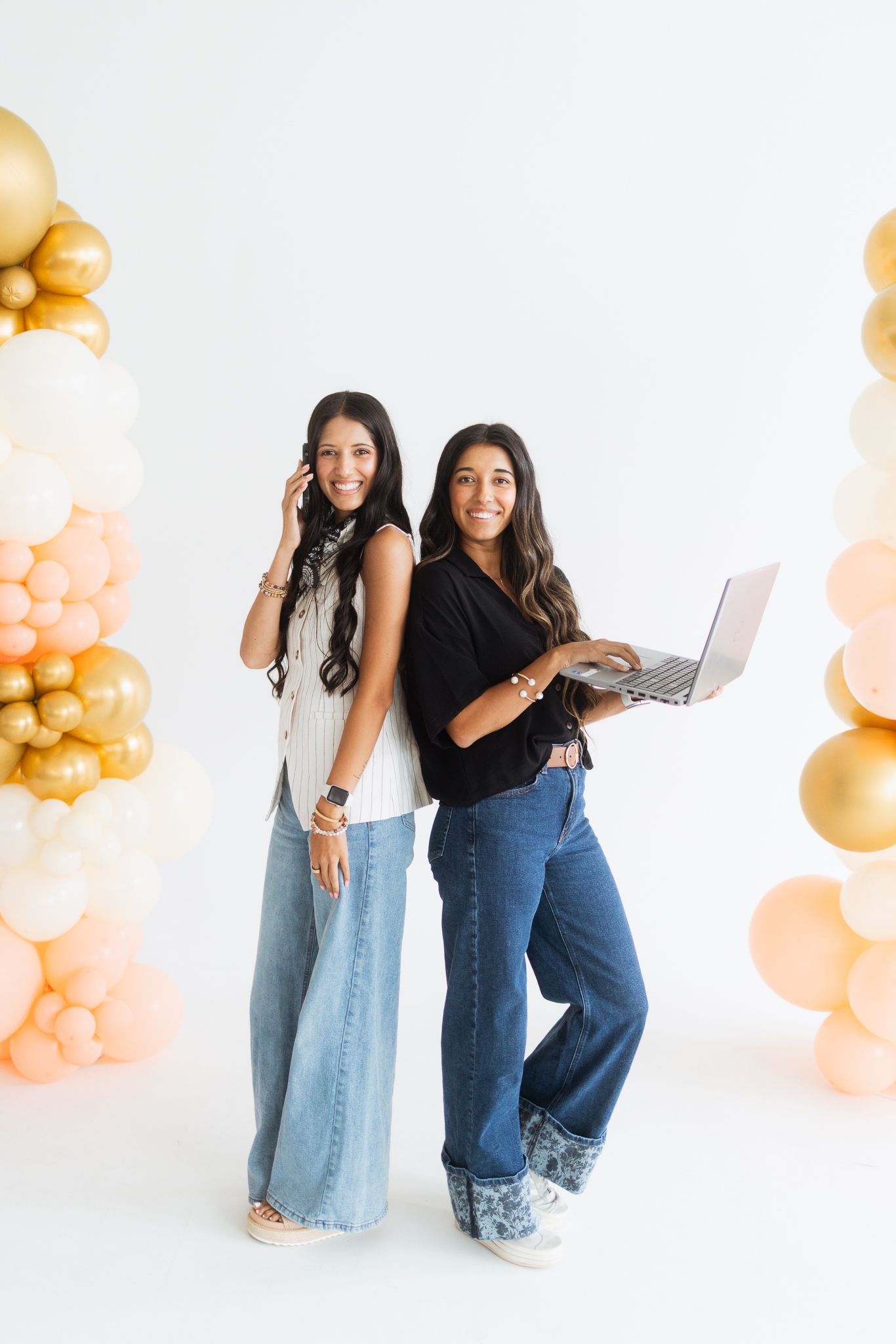 Two stylish women pose back-to-back with laptop and phone, surrounded by gold and pastel balloon columns.