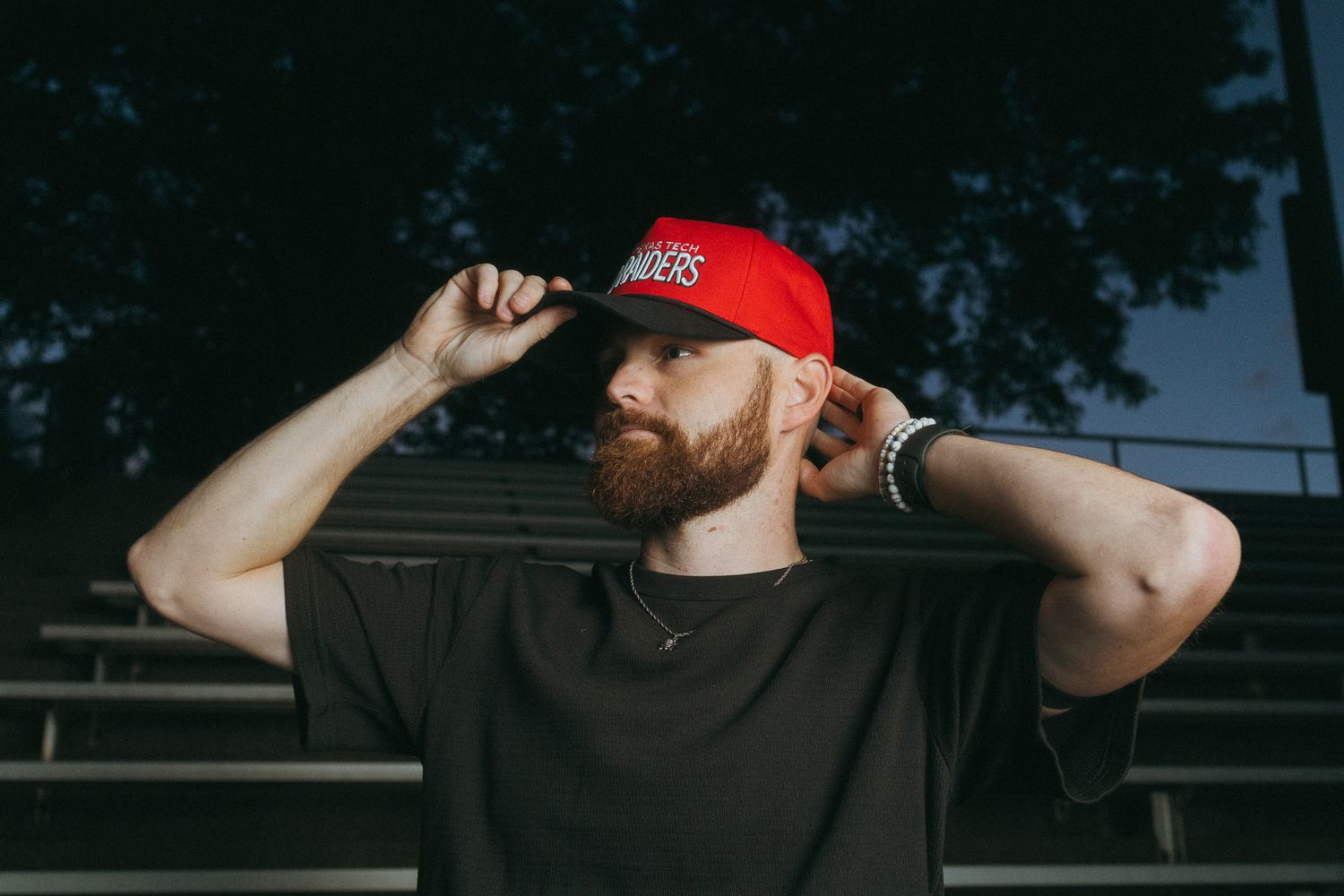 Bearded man adjusting a red snapback cap while standing in stadium bleachers.