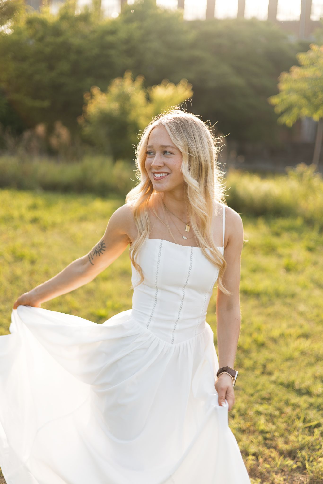 Smiling blonde woman in white sundress twirling in sunlit green field during golden hour.