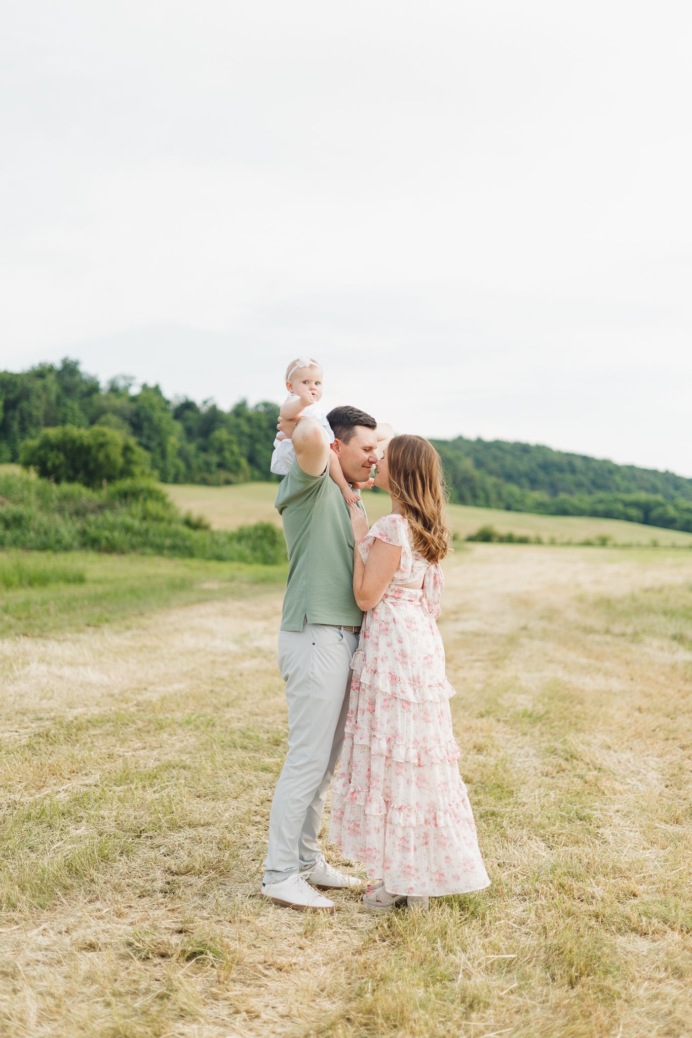 Happy young couple standing in a sunny field, man lifting baby girl while woman in floral dress looks on.