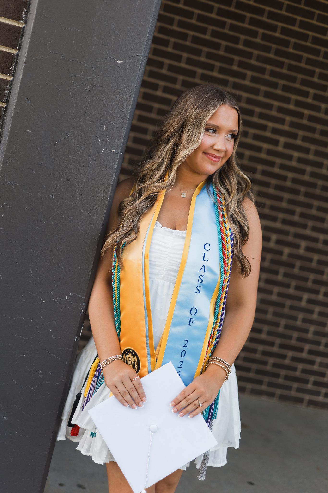 Graduating senior in white dress wears Class of 2023 sash and honor cords outside brick building.