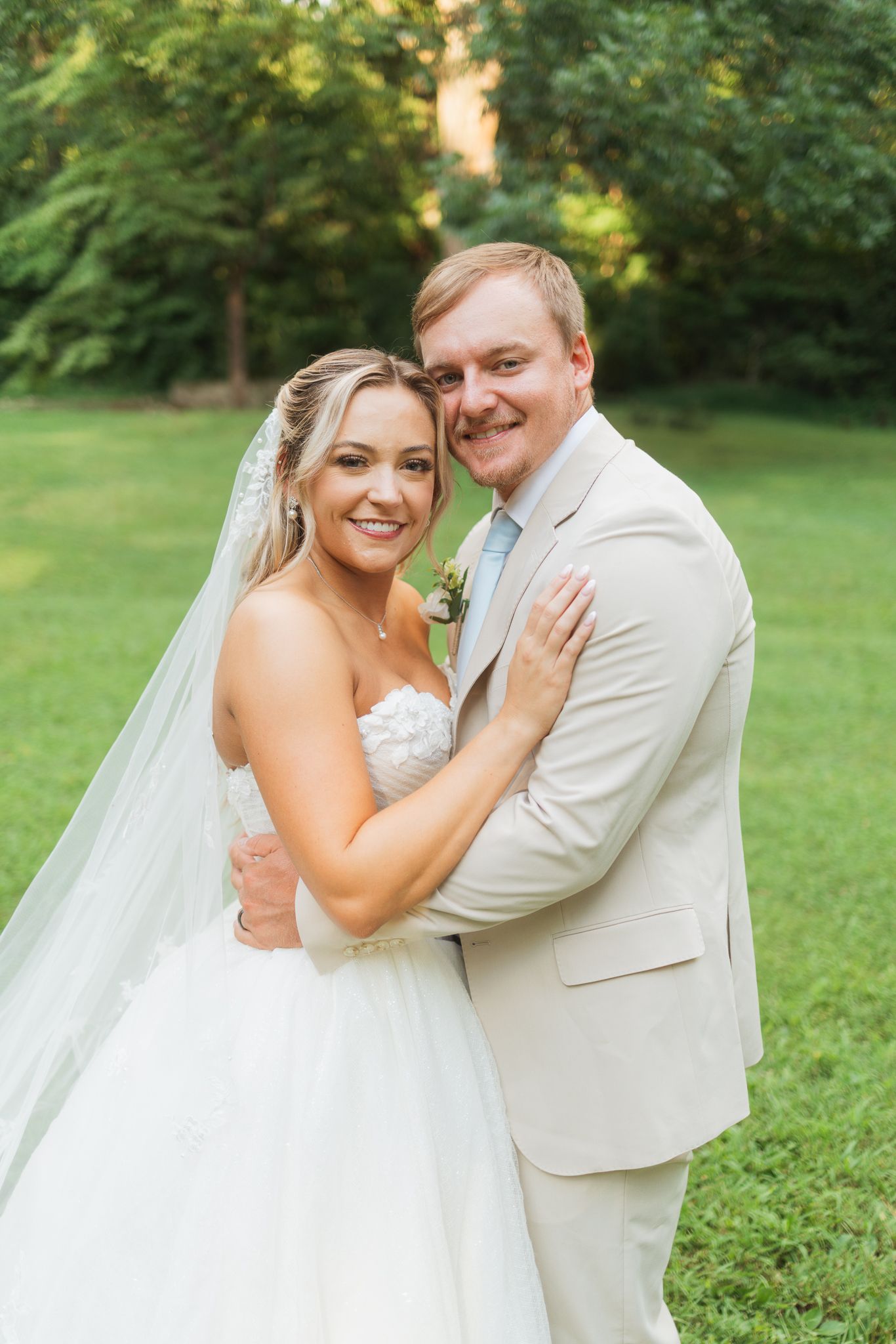 Smiling bride in strapless gown with veil embraces groom in tan suit outdoors on lush green lawn.