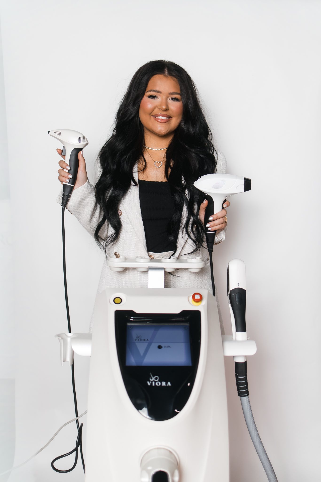 Smiling woman with long black hair holding two handpieces beside Viora laser aesthetic device.