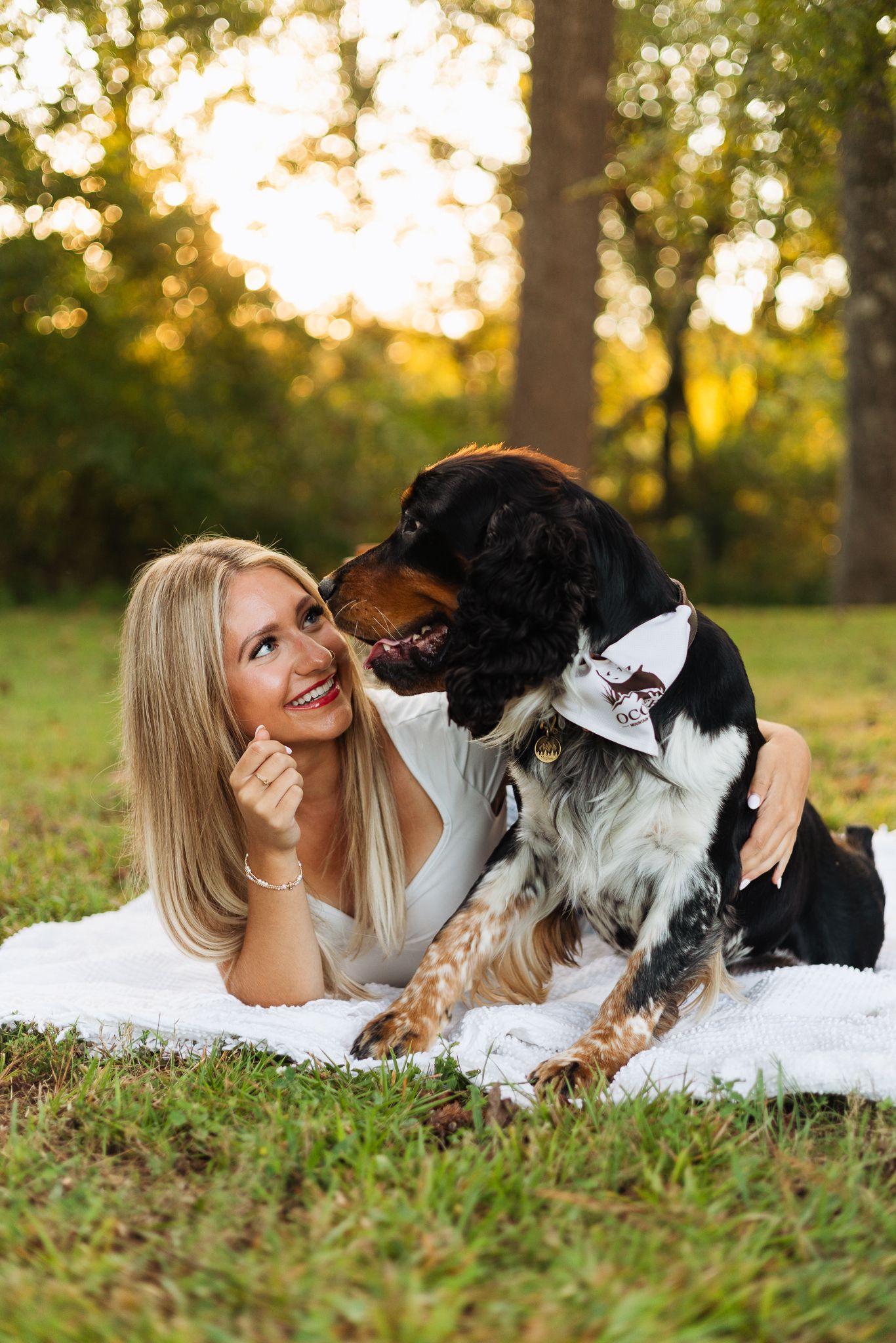 Smiling blonde woman lying on blanket with a tricolor Spaniel dog in sunny park.