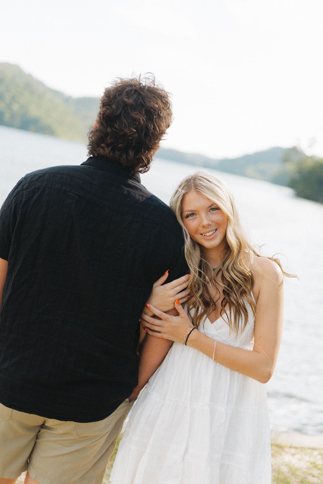 A smiling blonde woman in a white dress embraces a man in a black shirt near a scenic lake and mountains.