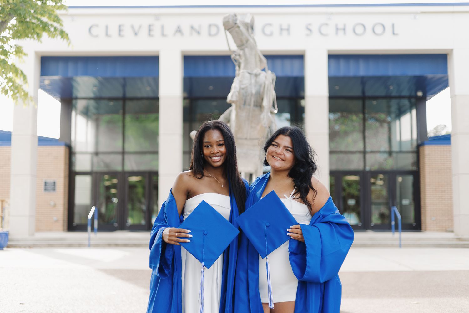 Two female graduates in blue caps and gowns posing in front of Cleveland High School.