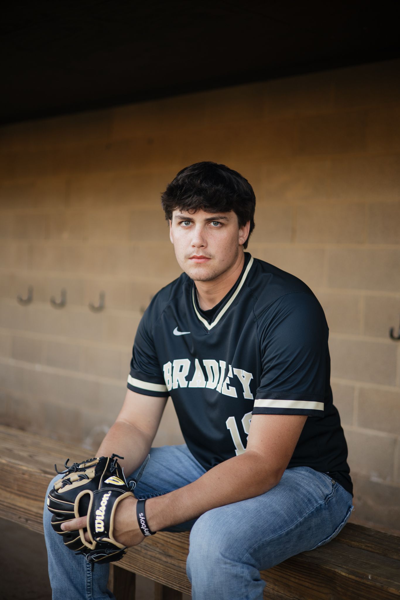 Young baseball player in Bradley jersey sitting in dugout holding Wilson glove, senior portrait photo.