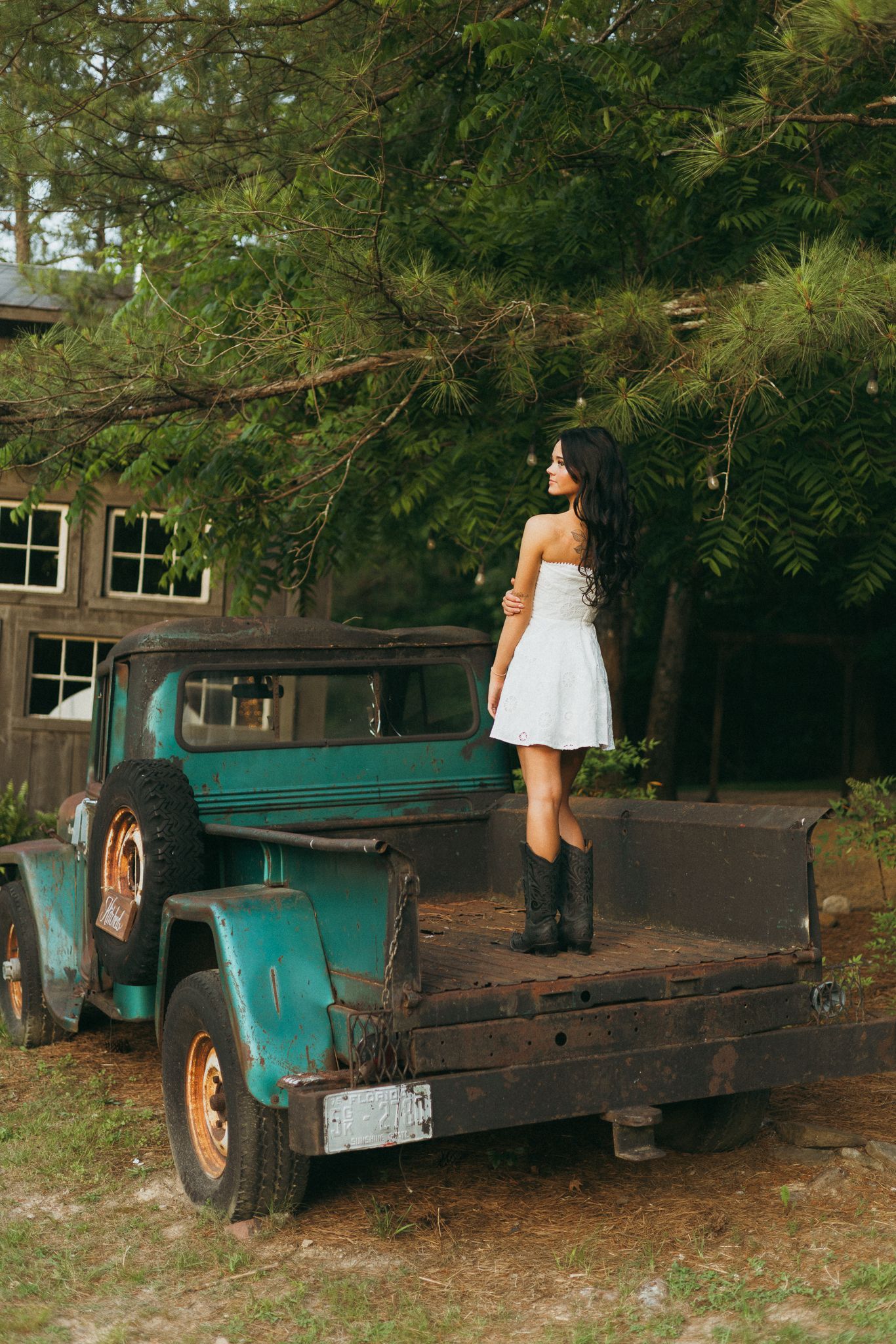 Young woman in white dress and cowboy boots standing on vintage rustic green truck bed.