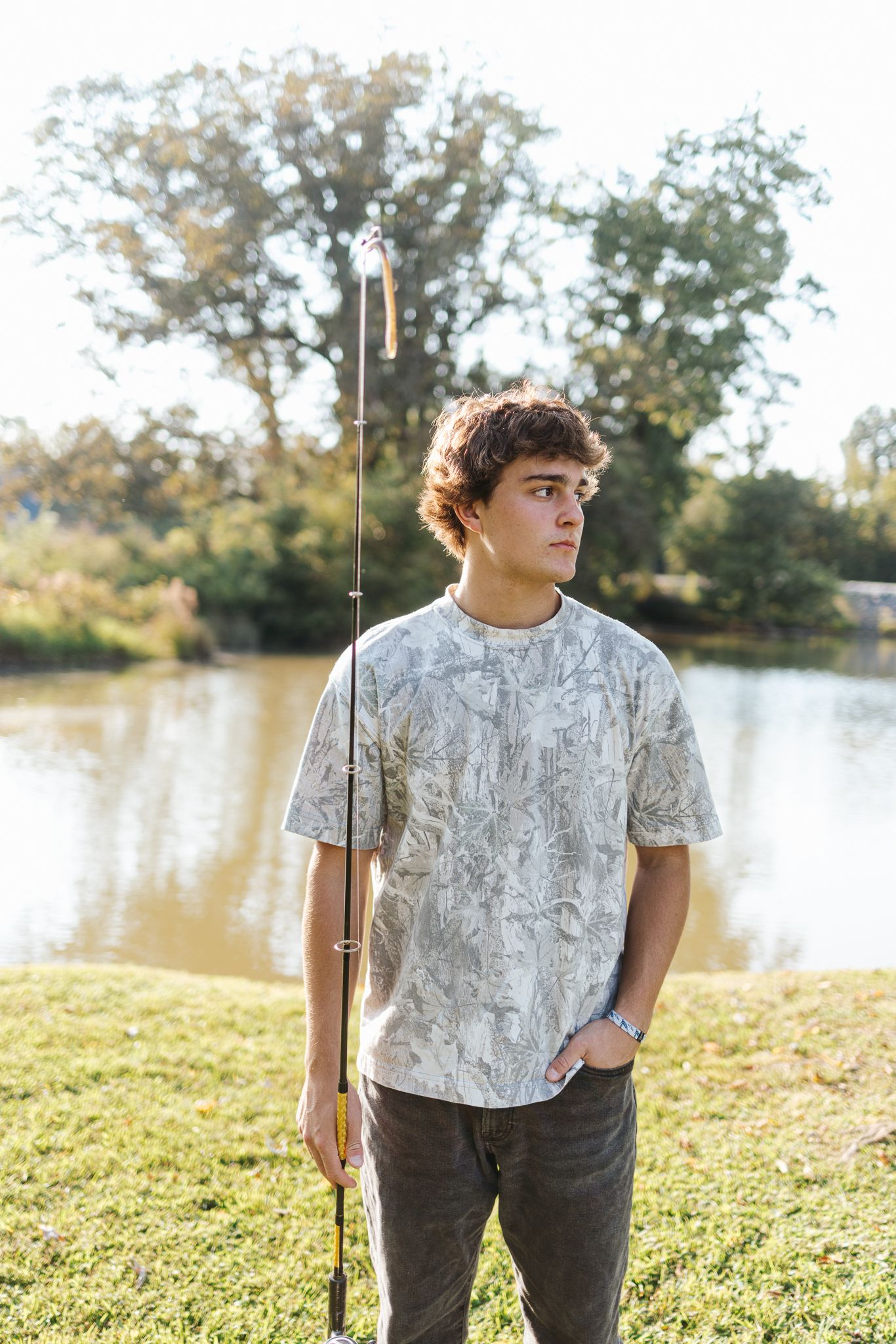 Young man holding fishing rod standing by a lake in a patterned t-shirt outdoors.
