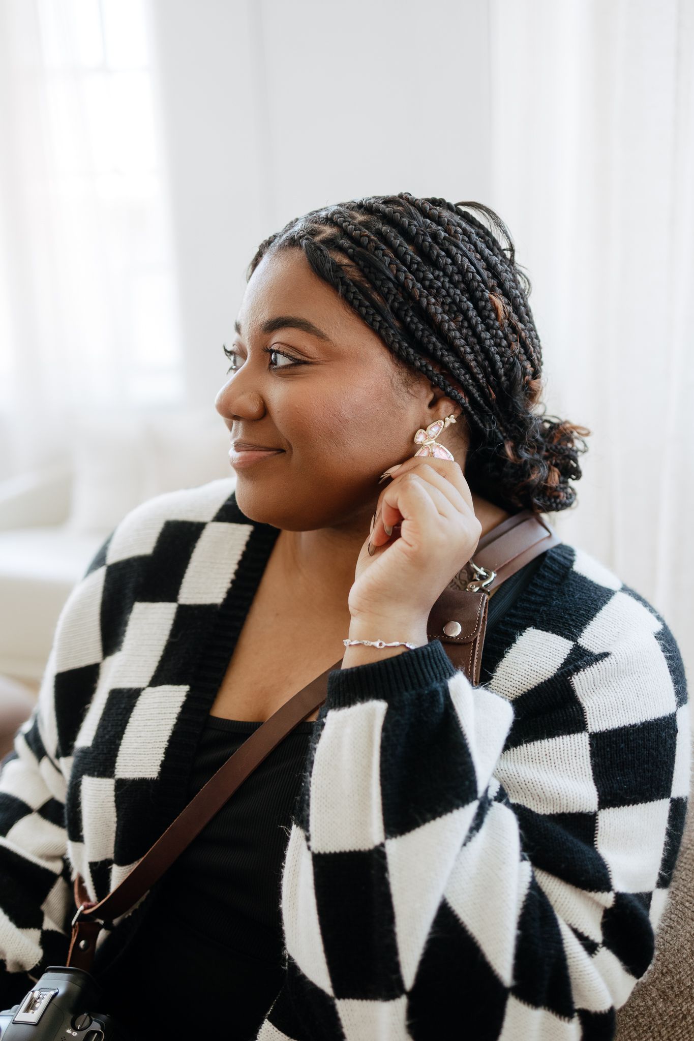Young Black woman with braids adjusting earring wearing black and white checkered cardigan.