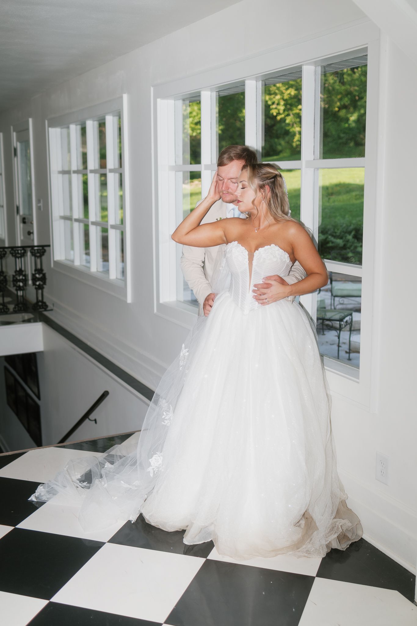 Bride in strapless white ballgown embraces groom near large windows on black and white checkered floor.
