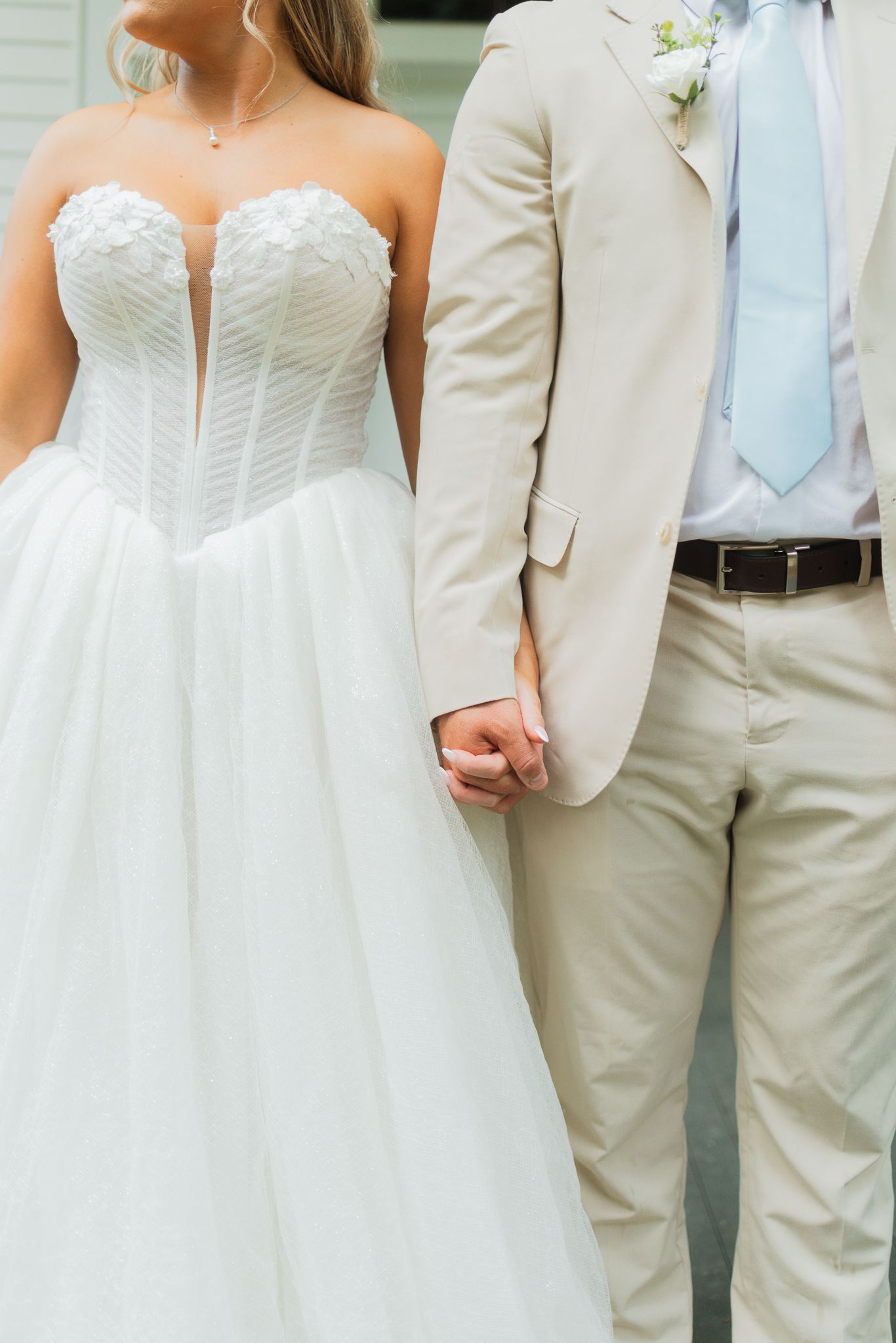Bride in strapless white corset wedding gown holds hands with groom in beige suit and light blue tie.