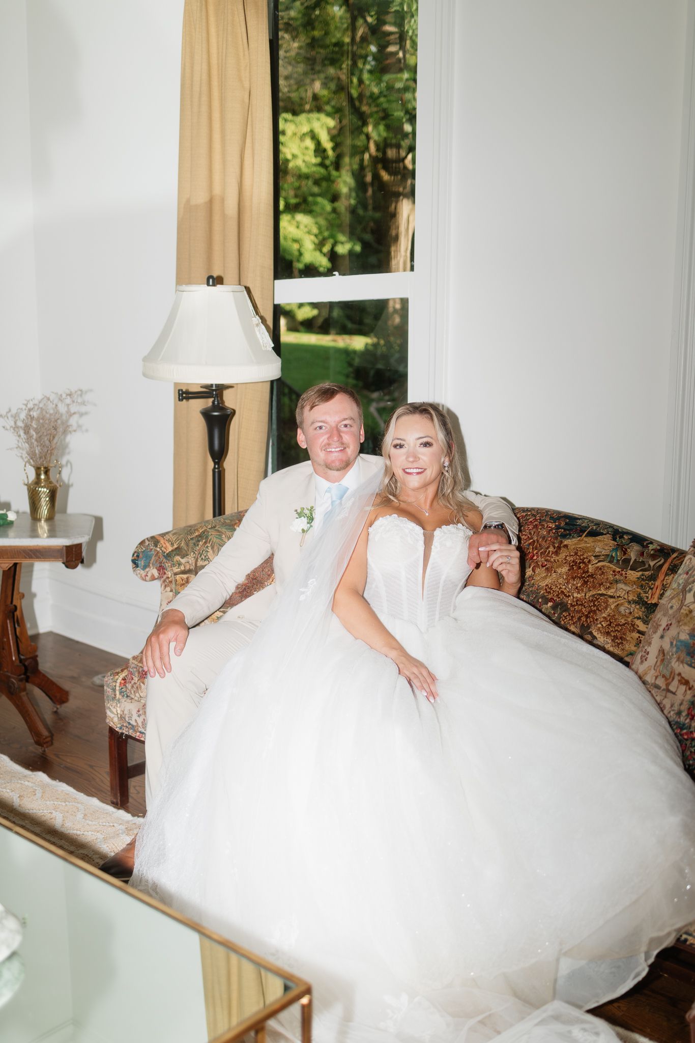 Bride and groom sitting together on ornate sofa in elegant white room, bride wearing full ballgown wedding dress.