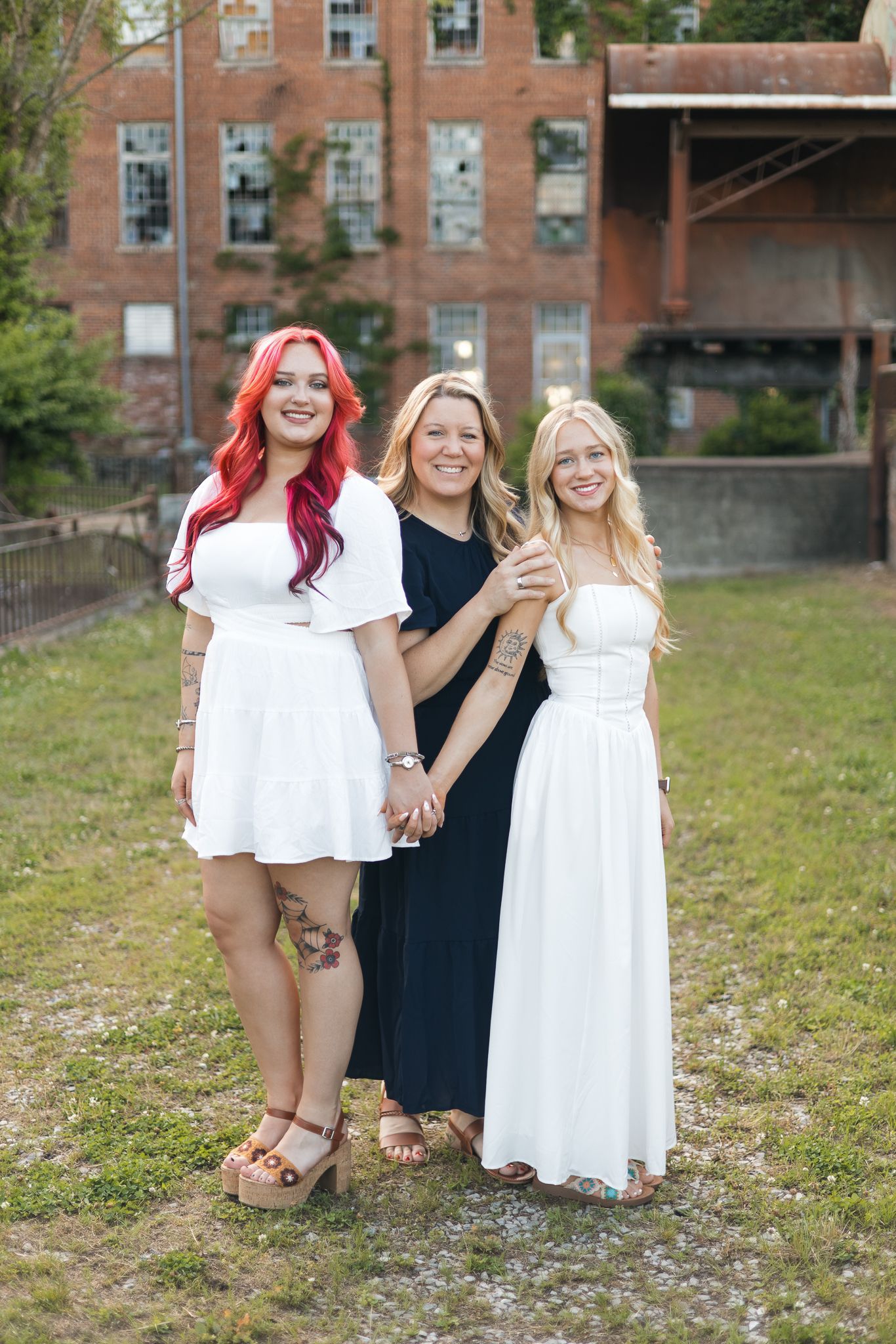 Three women in white and black dresses pose together outdoors near a historic brick building.