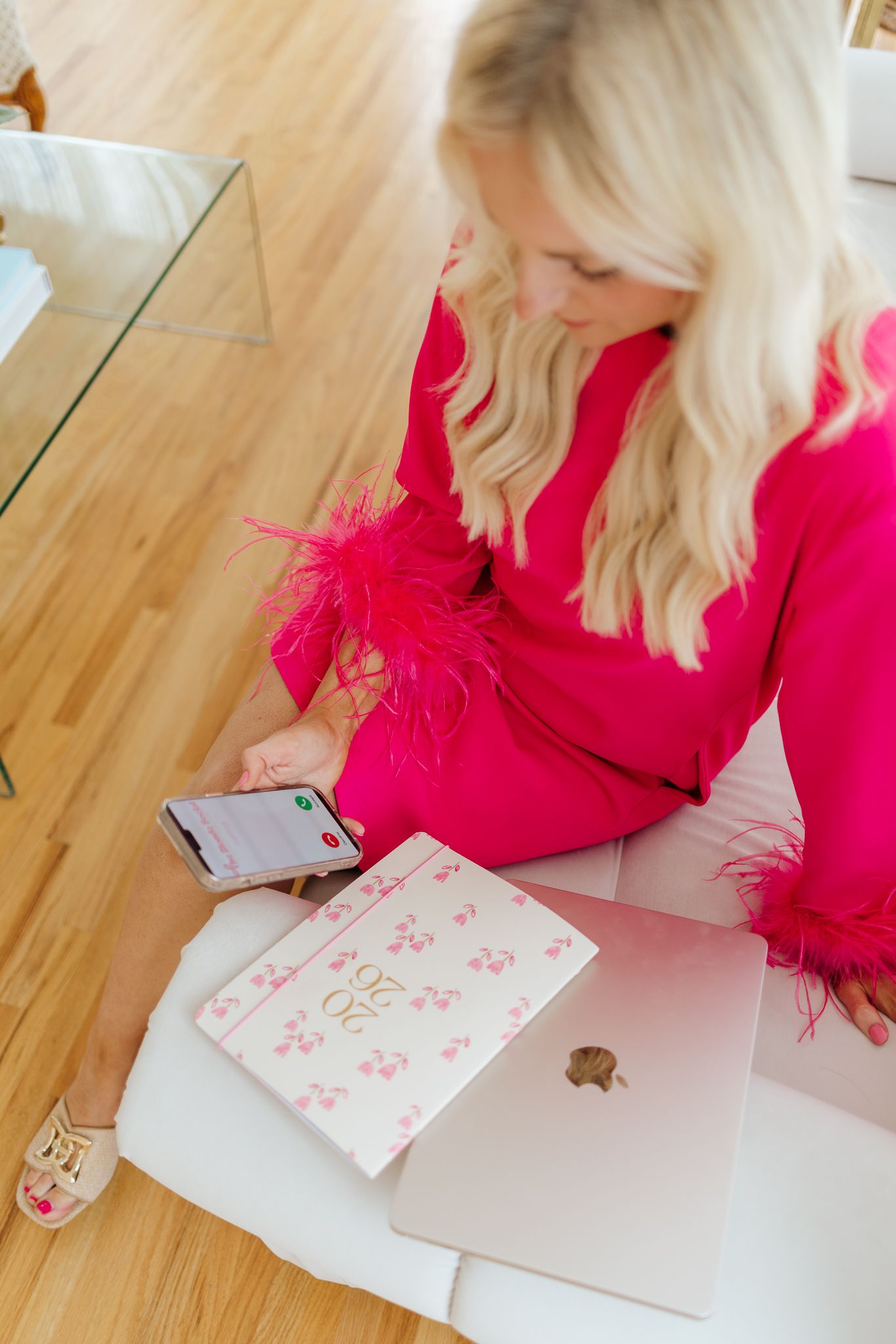Overhead view of blonde woman in pink feather top using smartphone beside laptop and planner.