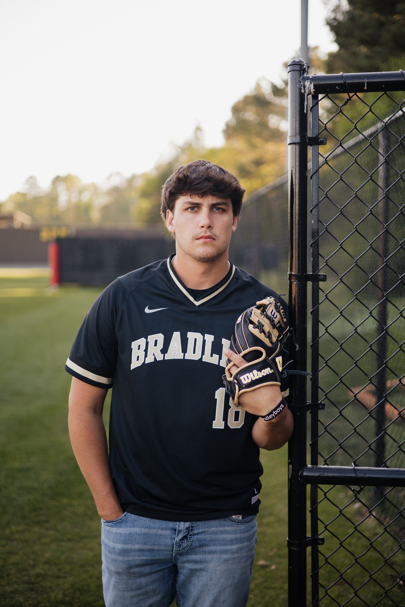Young baseball player in Bradley jersey #18 holding a Wilson glove, leaning against a chain-link fence on a field.