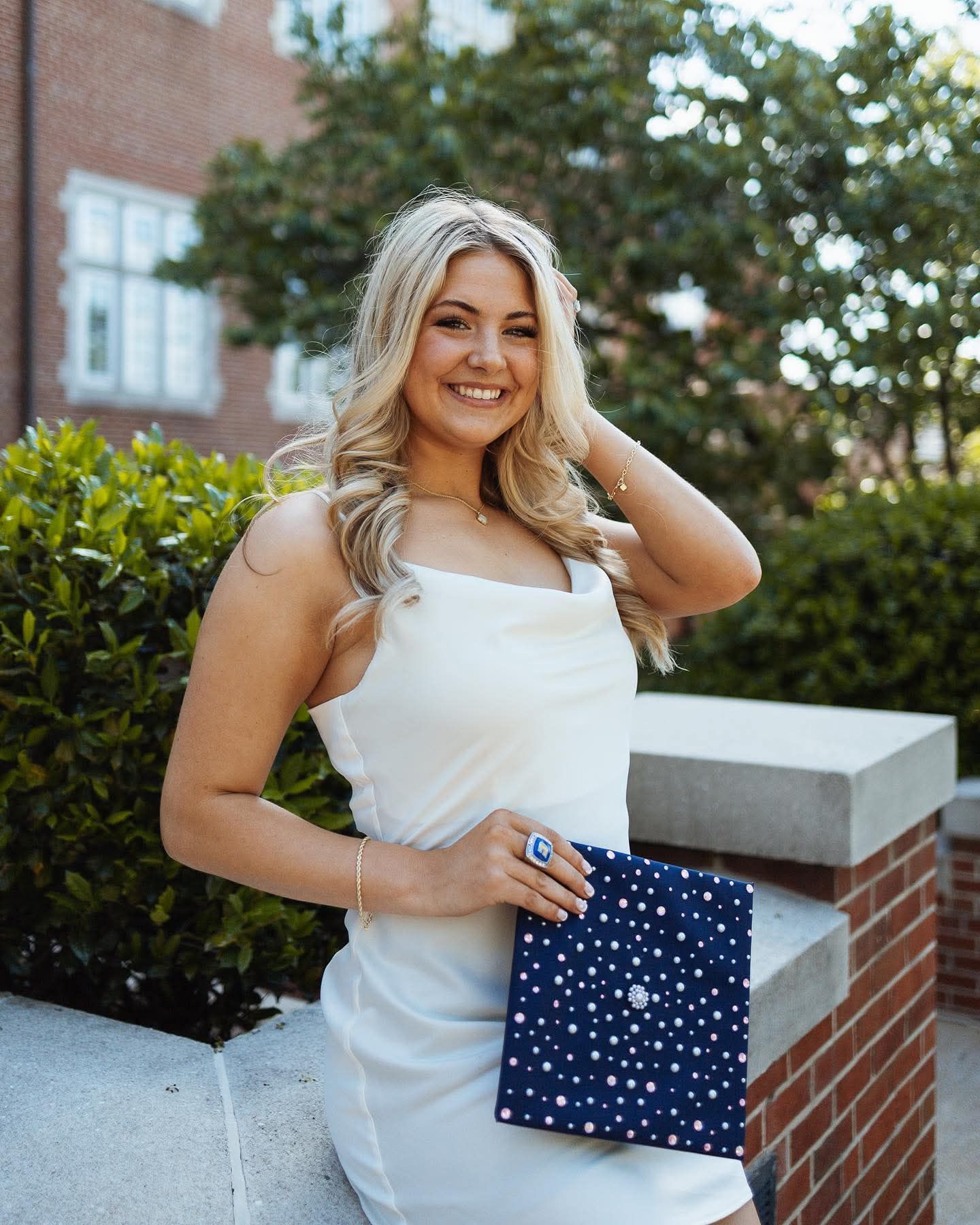 Smiling blonde woman in white dress holding navy blue graduation cap with white stars outdoors.