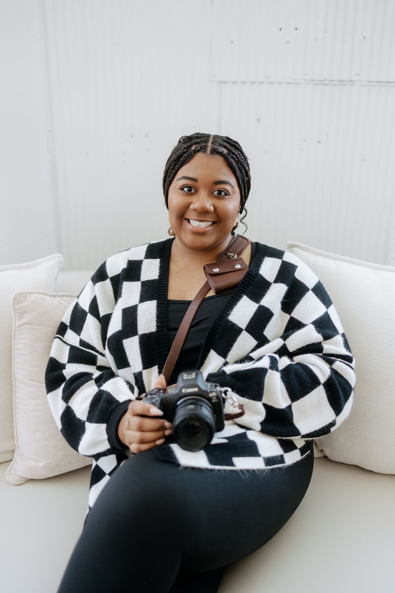 Cheerful Black female photographer holding camera on white sofa in bright studio setting.