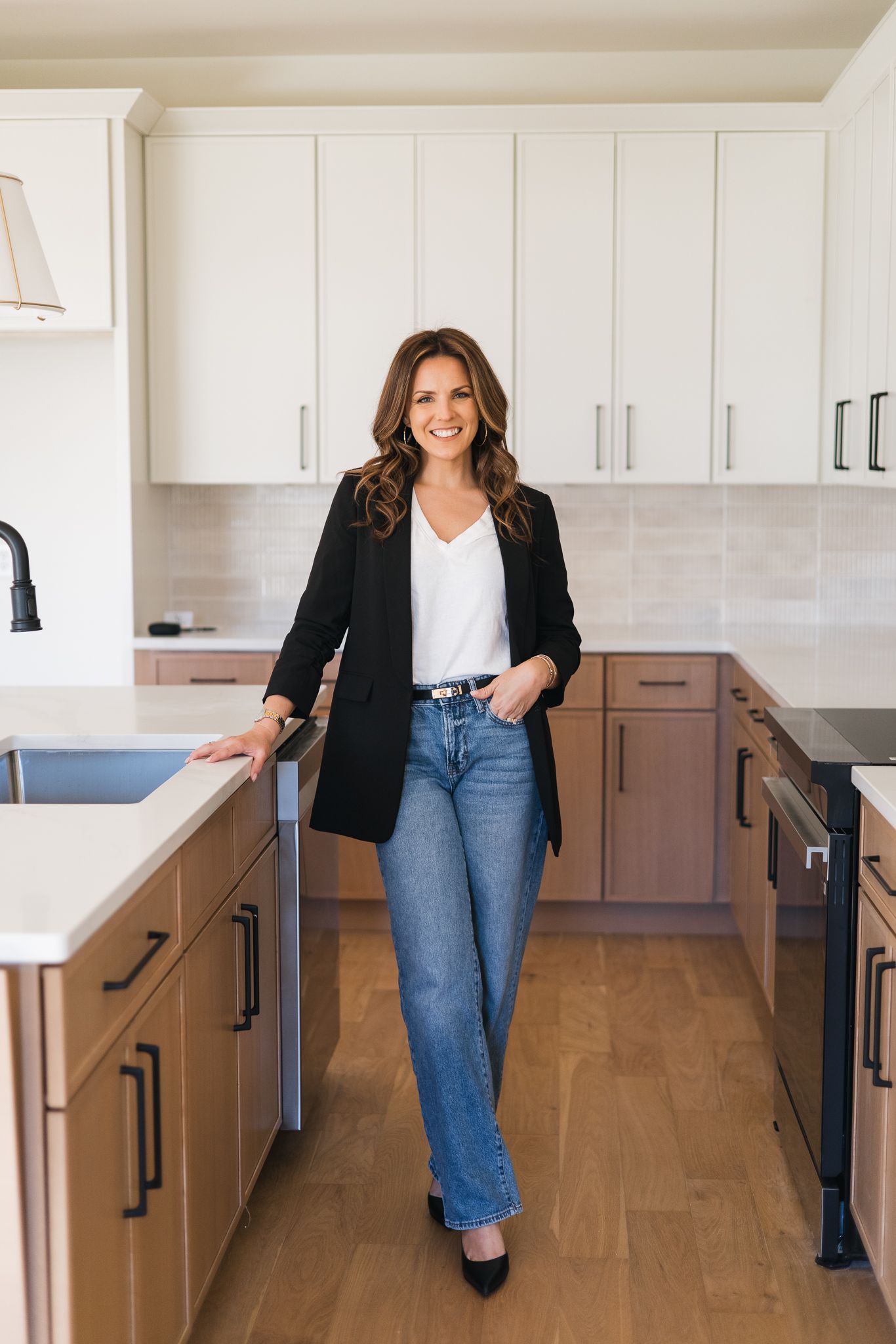 Brunette woman in black blazer and jeans leaning on kitchen counter in modern white kitchen.
