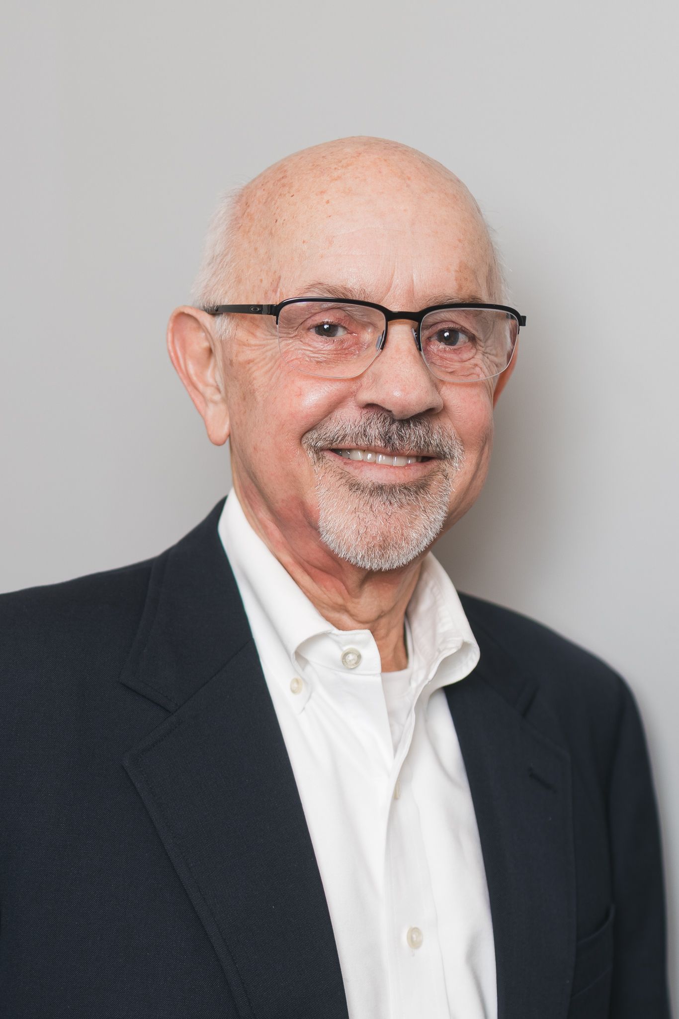 Smiling elderly bald man with glasses and gray goatee wearing a dark blazer and white shirt.