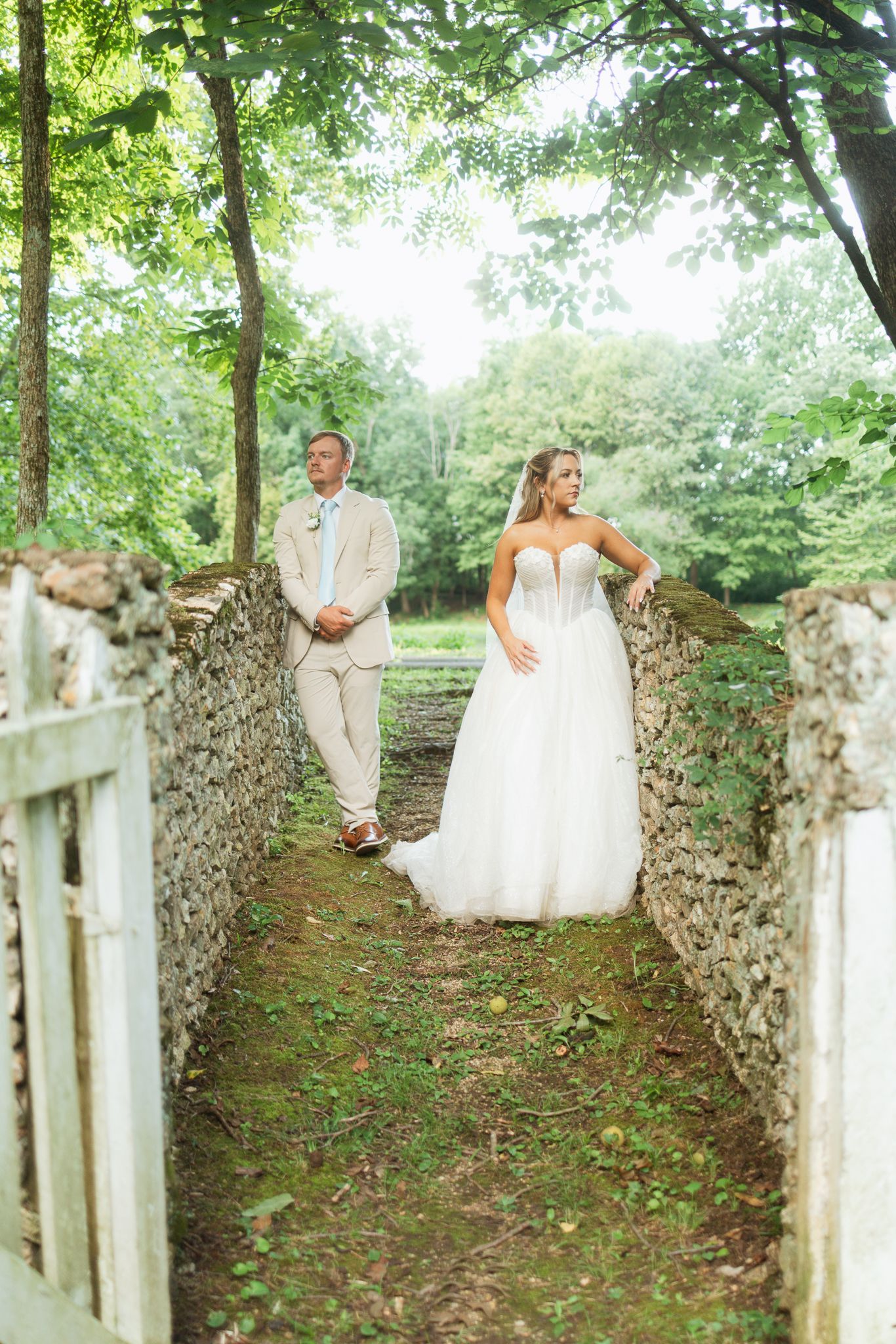 Bride in white gown and groom in tan suit walk through stone-walled garden path surrounded by lush green trees.