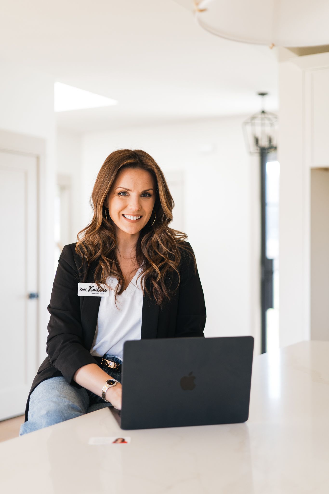 Smiling brunette woman in black blazer working on MacBook laptop at white kitchen counter.