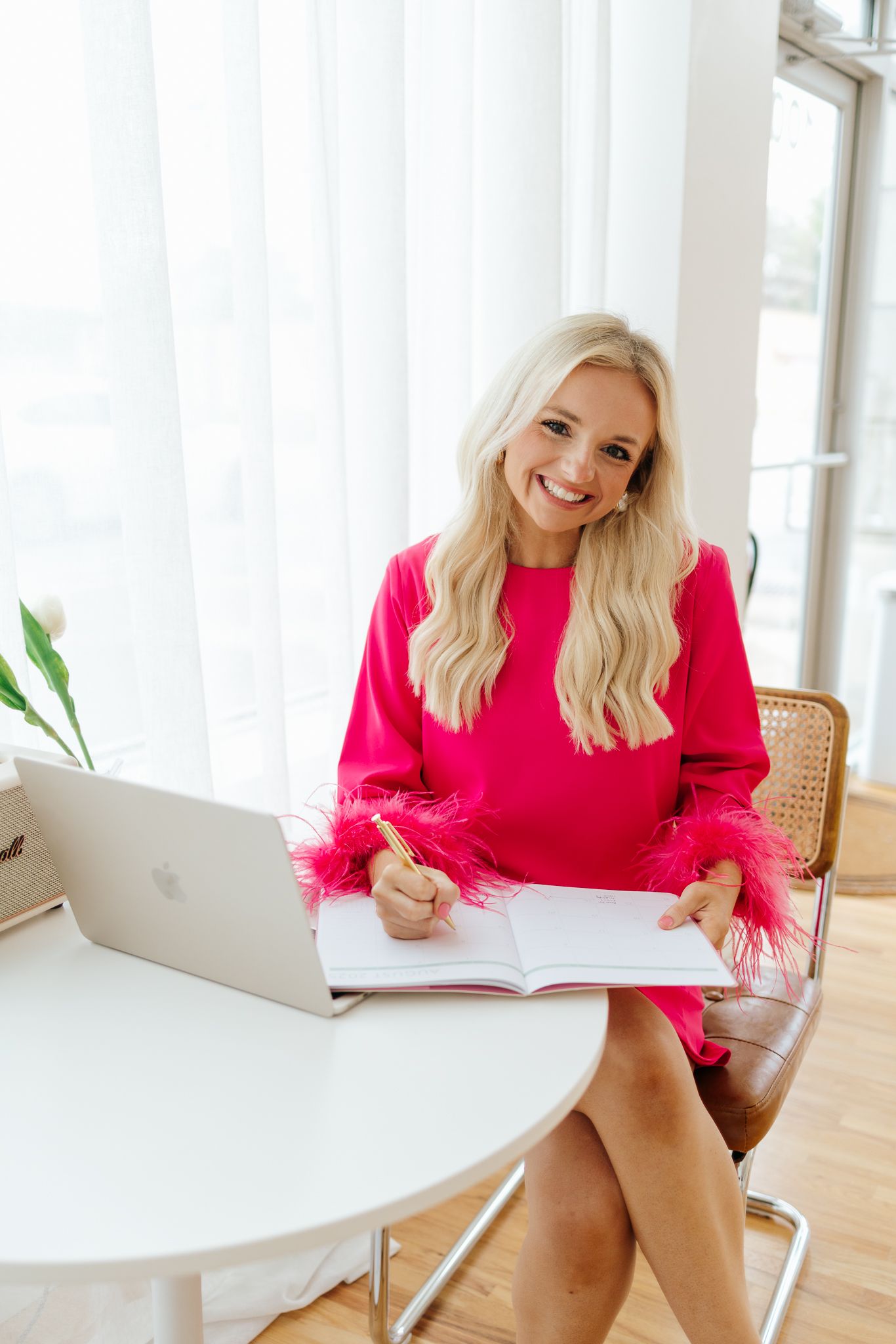 Smiling blonde woman in pink feather dress writes in notebook at desk with open laptop.