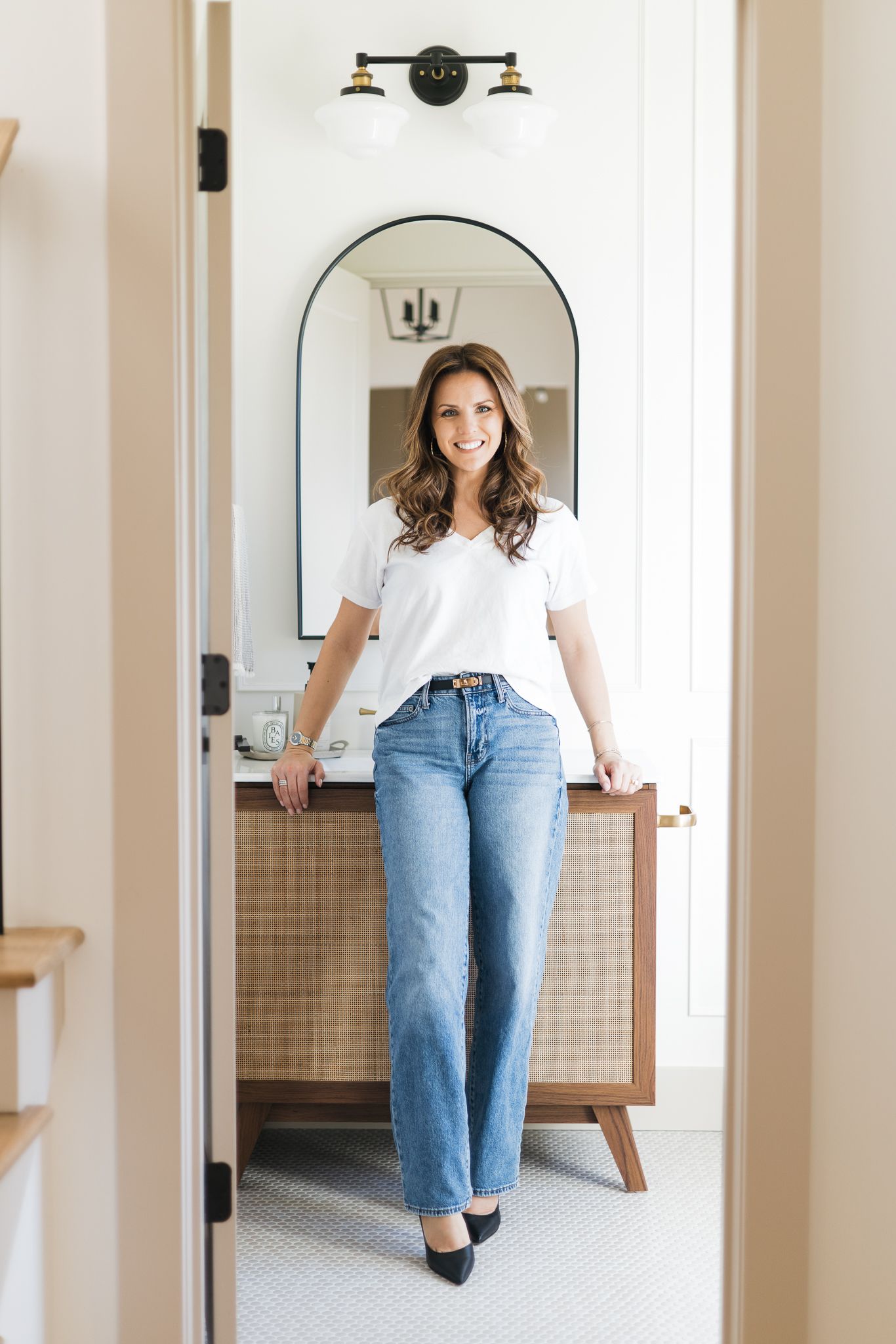 Smiling brunette woman in white tee and wide-leg jeans standing in doorway of stylish modern bathroom.